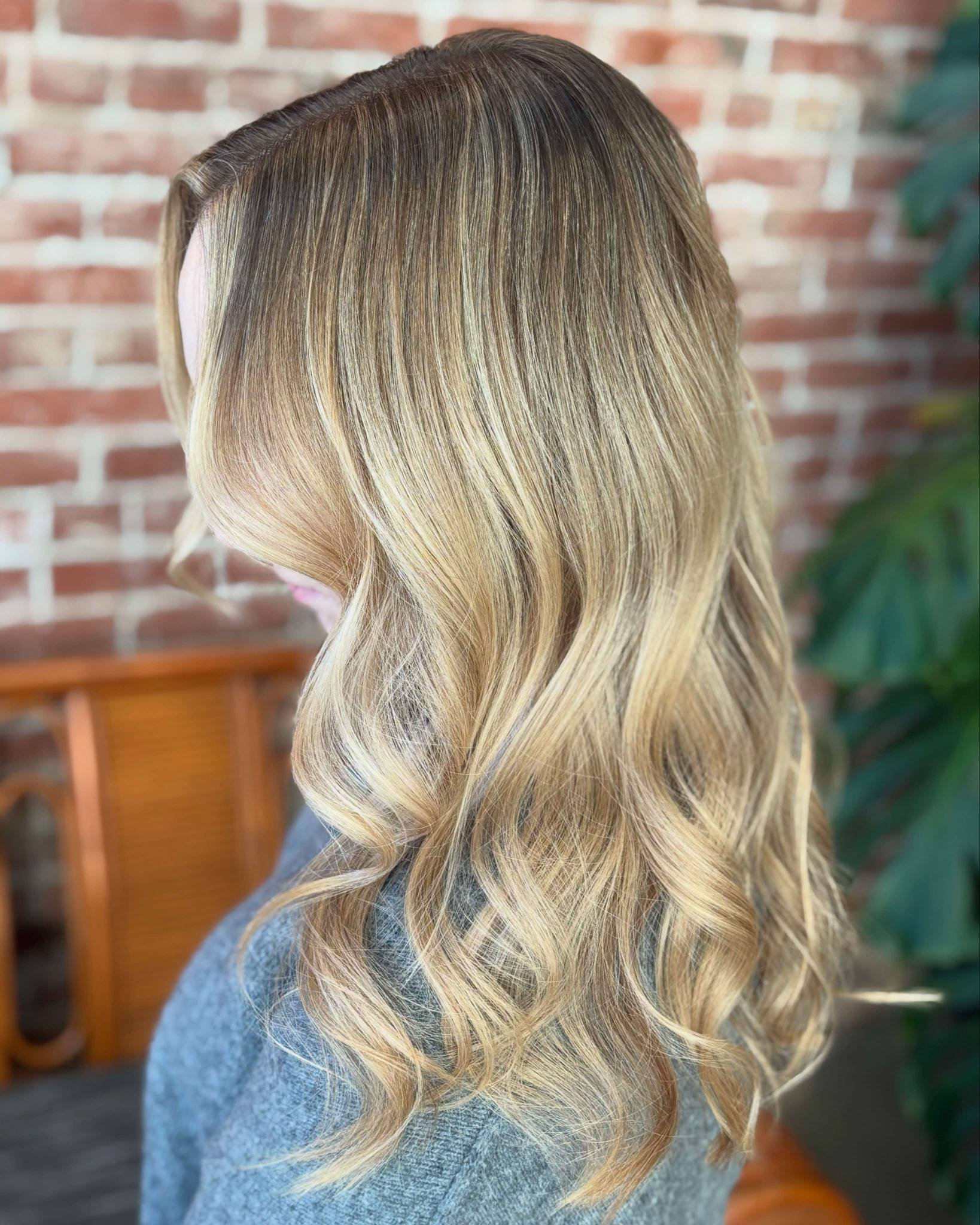 Close-up of a woman's blonde hair styled in loose waves, with a background featuring a brick wall and greenery.