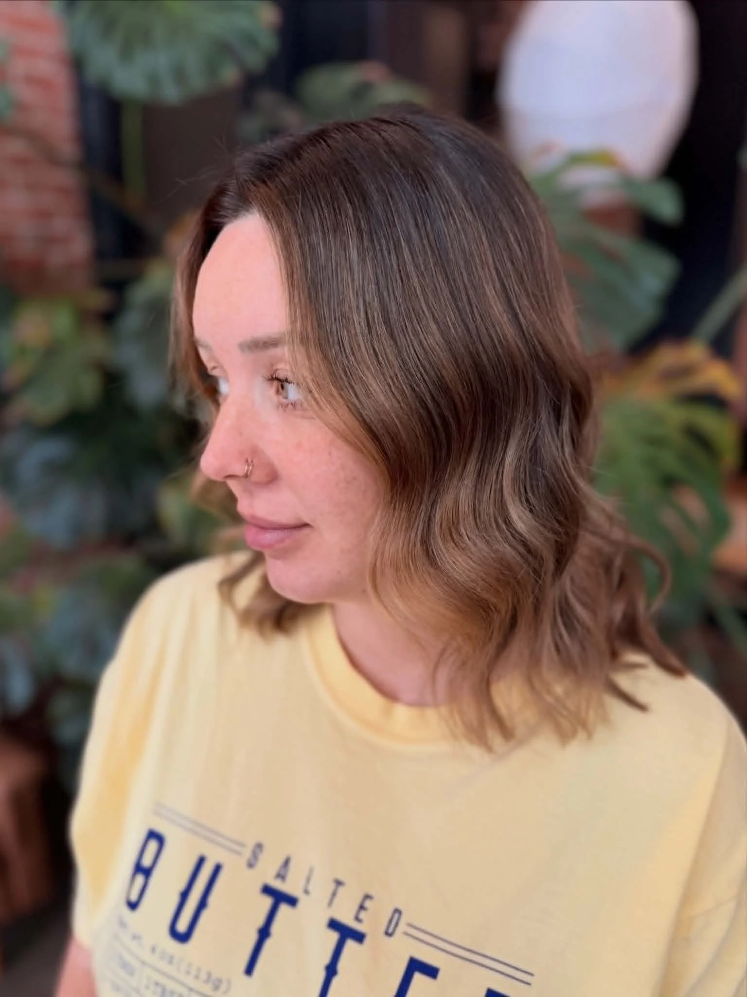 Close-up of a woman with shoulder-length brown hair, wearing a beige shirt, standing indoors with plants and a brick wall in the background.