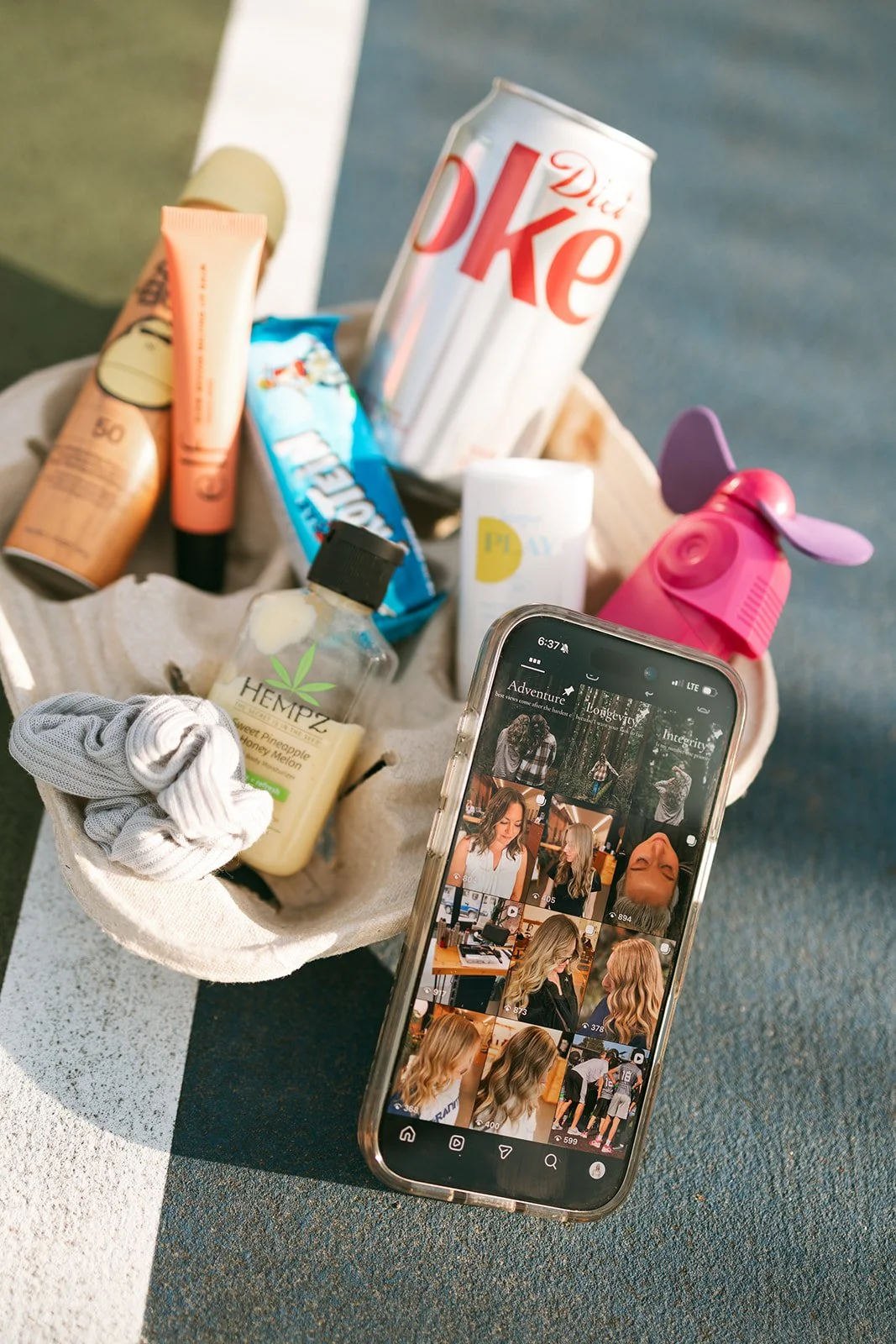 A picnic basket filled with various items including a can of Diet Coke, a bottle of hemp lotion, a stick of deodorant, a face spray, a snack bar, a pink spray bottle, a pair of socks, and a white bottle, with a smartphone showing a collage of hair styling photos resting on the basket.