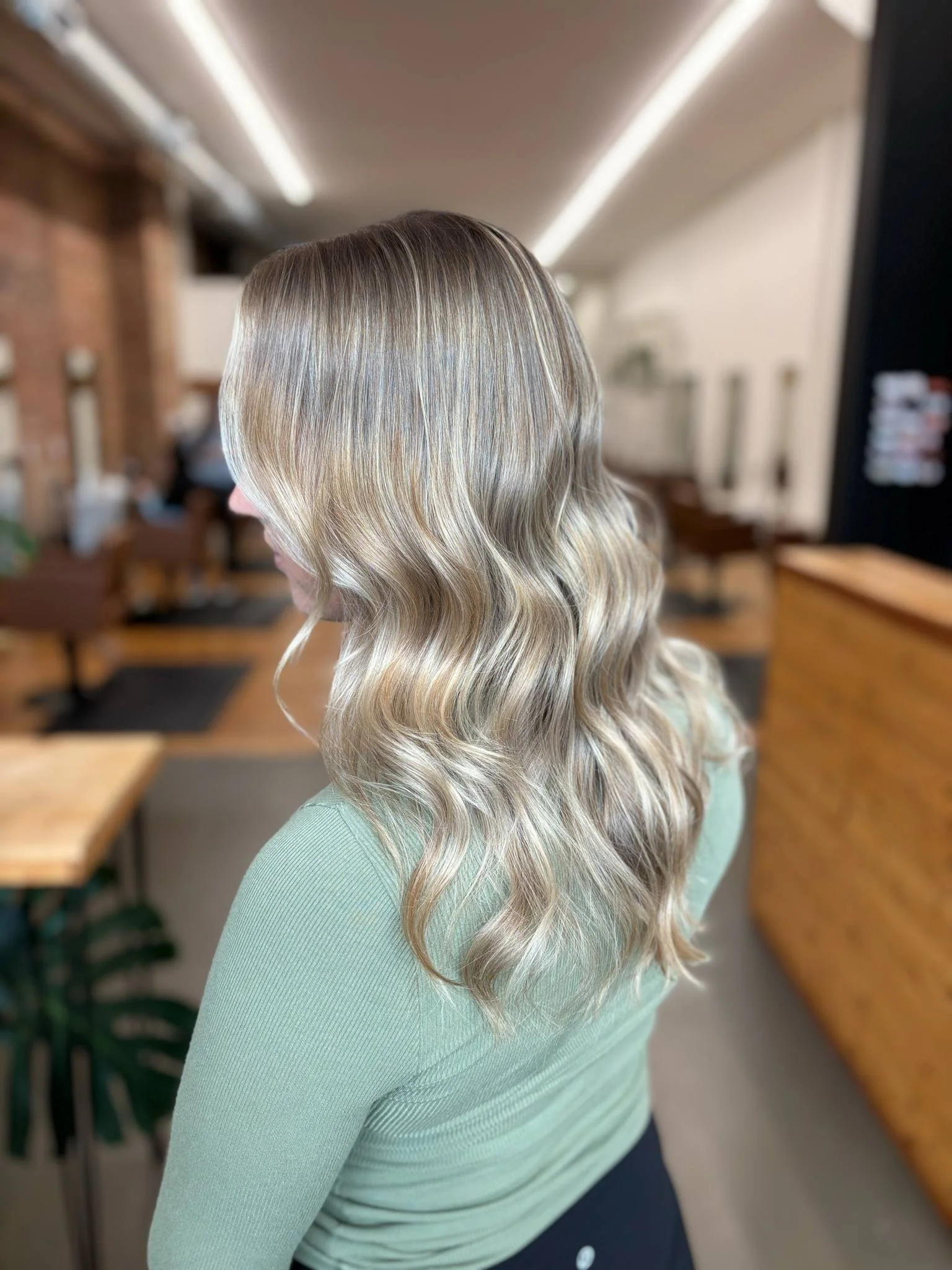 Woman with blonde, wavy hair wearing a light green top in a salon or similar indoor space.