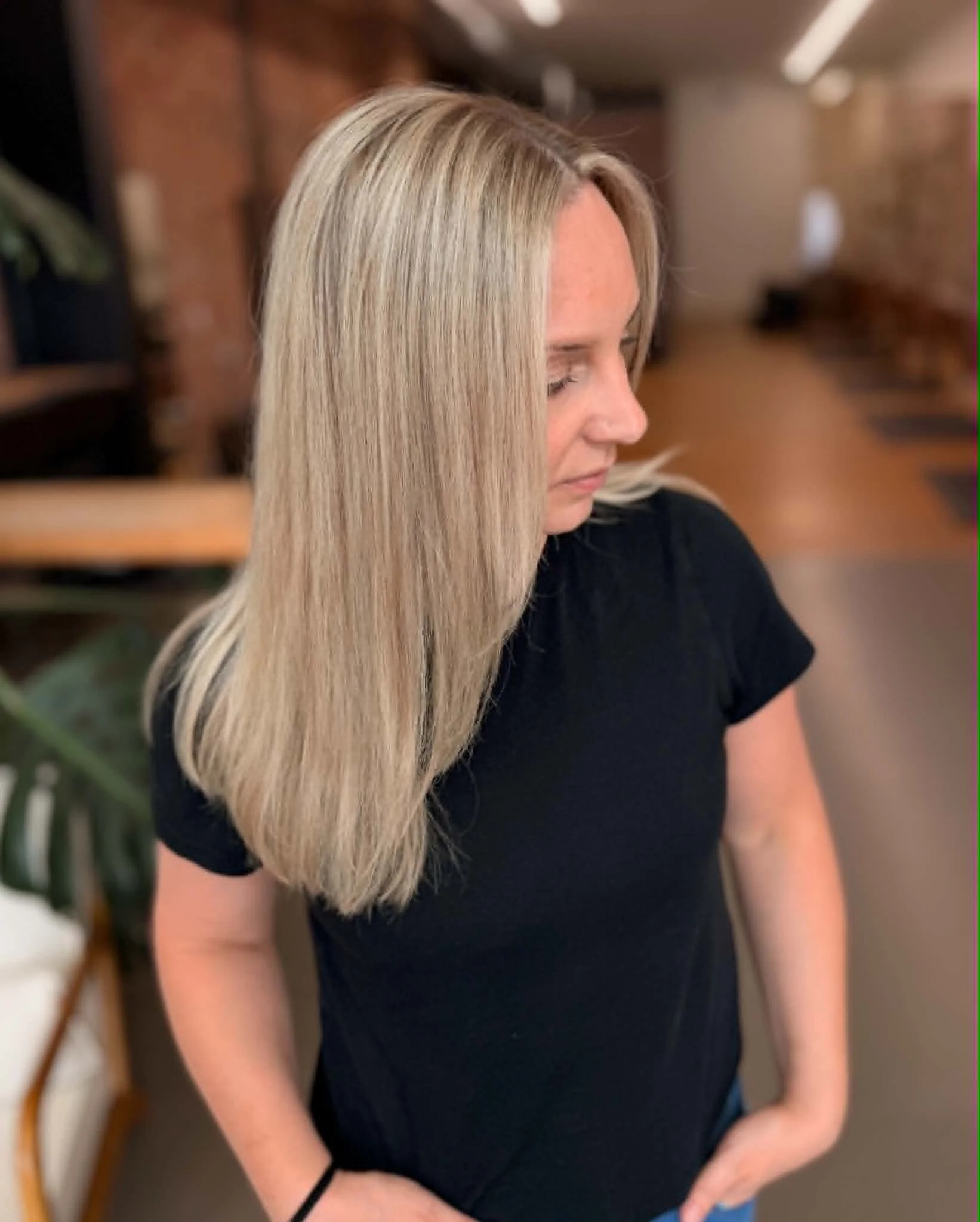 A woman with long, straight blonde hair in a salon or similar setting. She is wearing a black top and appears to be looking down or away. The background includes blurred brown brick walls and indoor lighting.