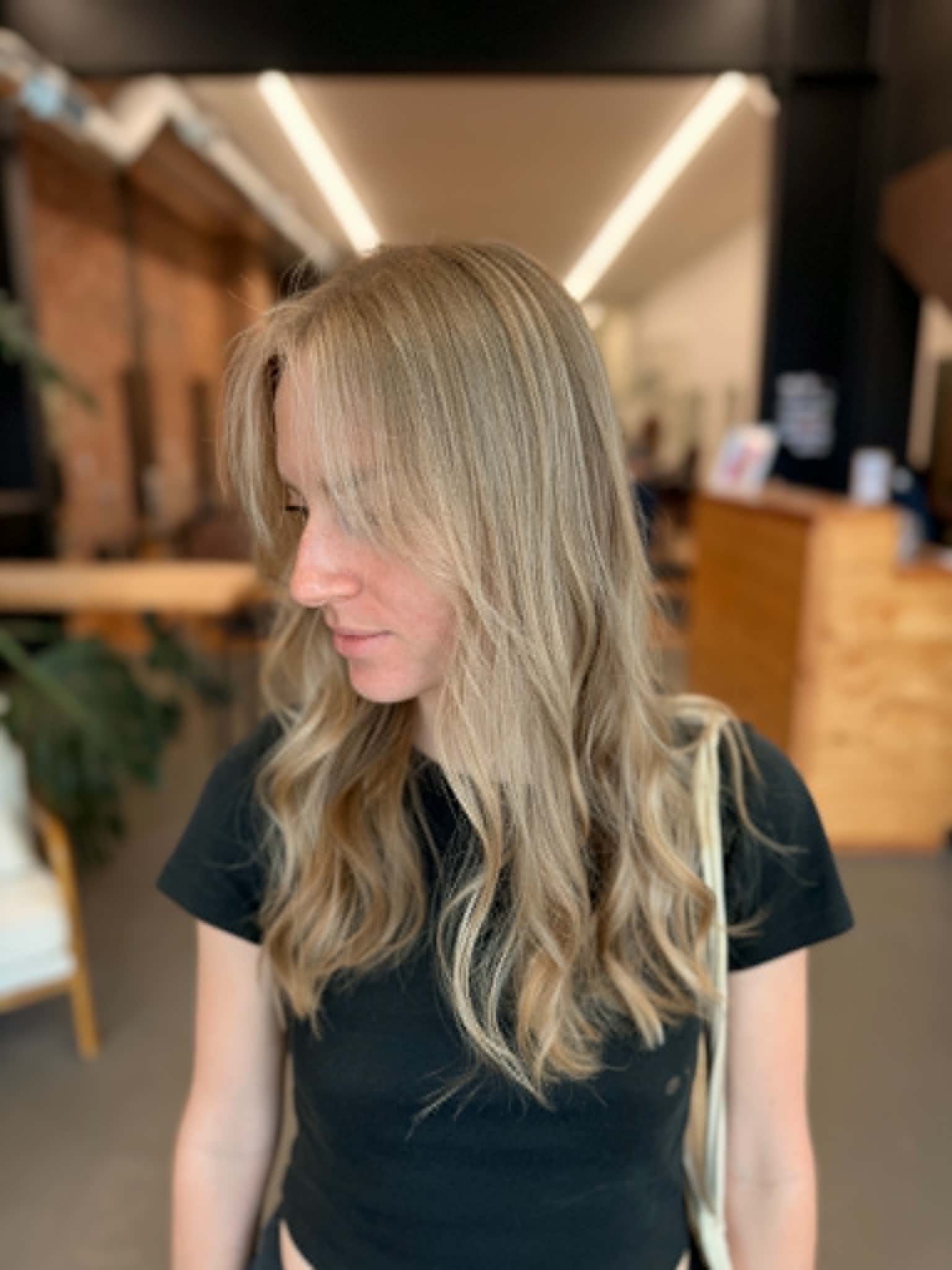 A woman with long, wavy blonde hair wearing a black t-shirt, standing in a retail store with wooden shelves and a mirror in the background.