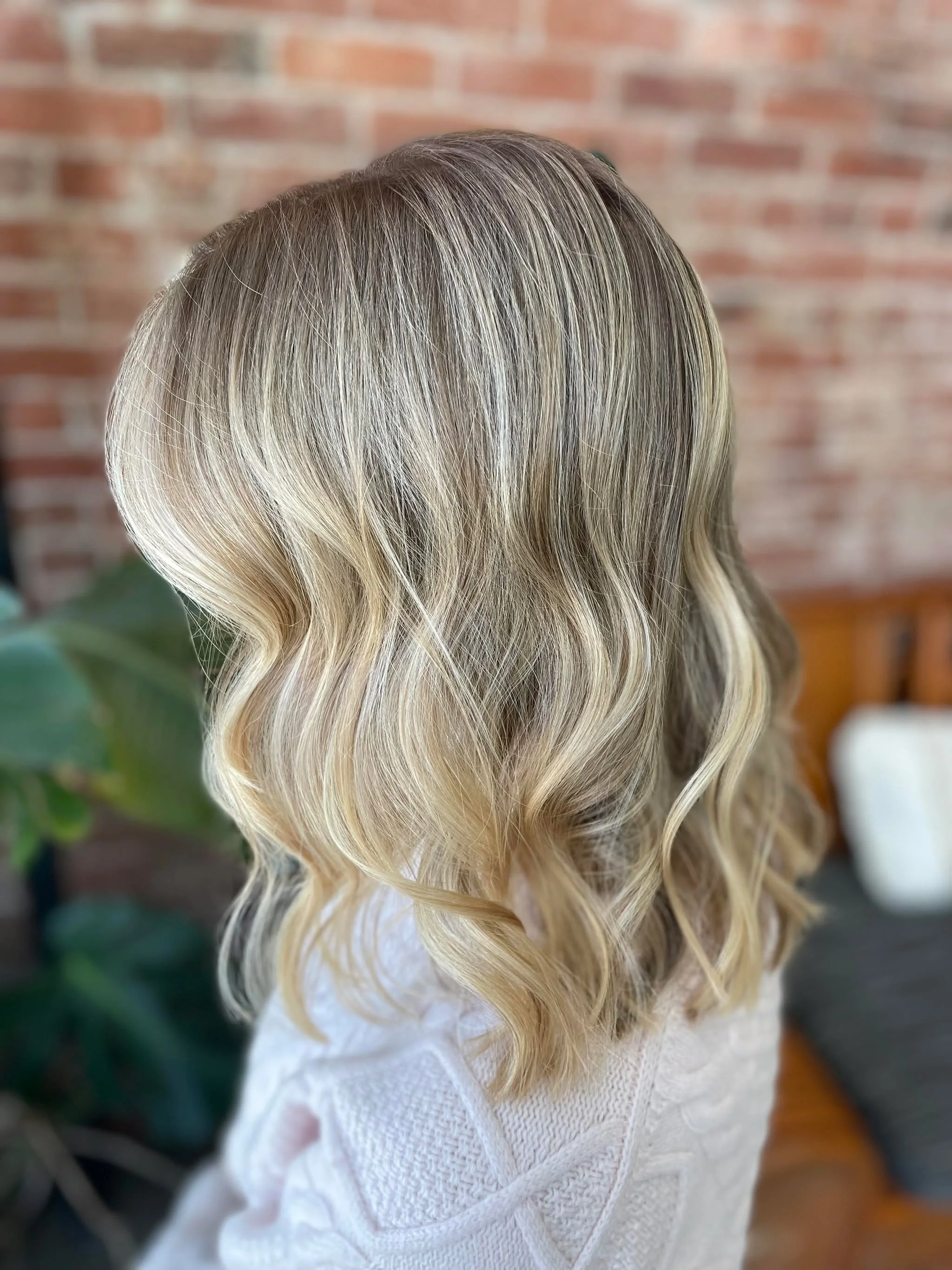 Woman with long, wavy, blonde hair standing indoors with a brick wall in the background.