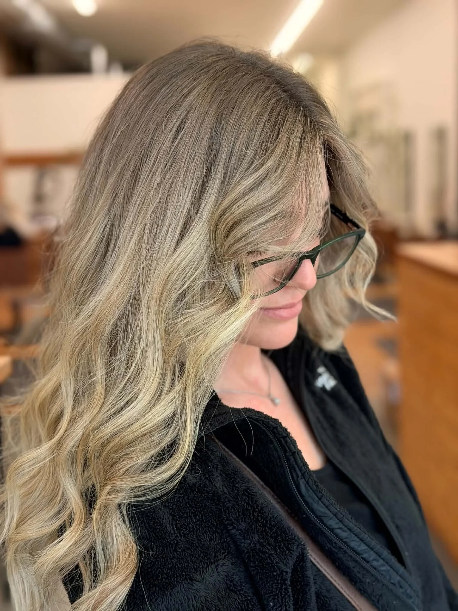 A woman with long, wavy blonde hair wearing glasses and a black jacket, looking downward.