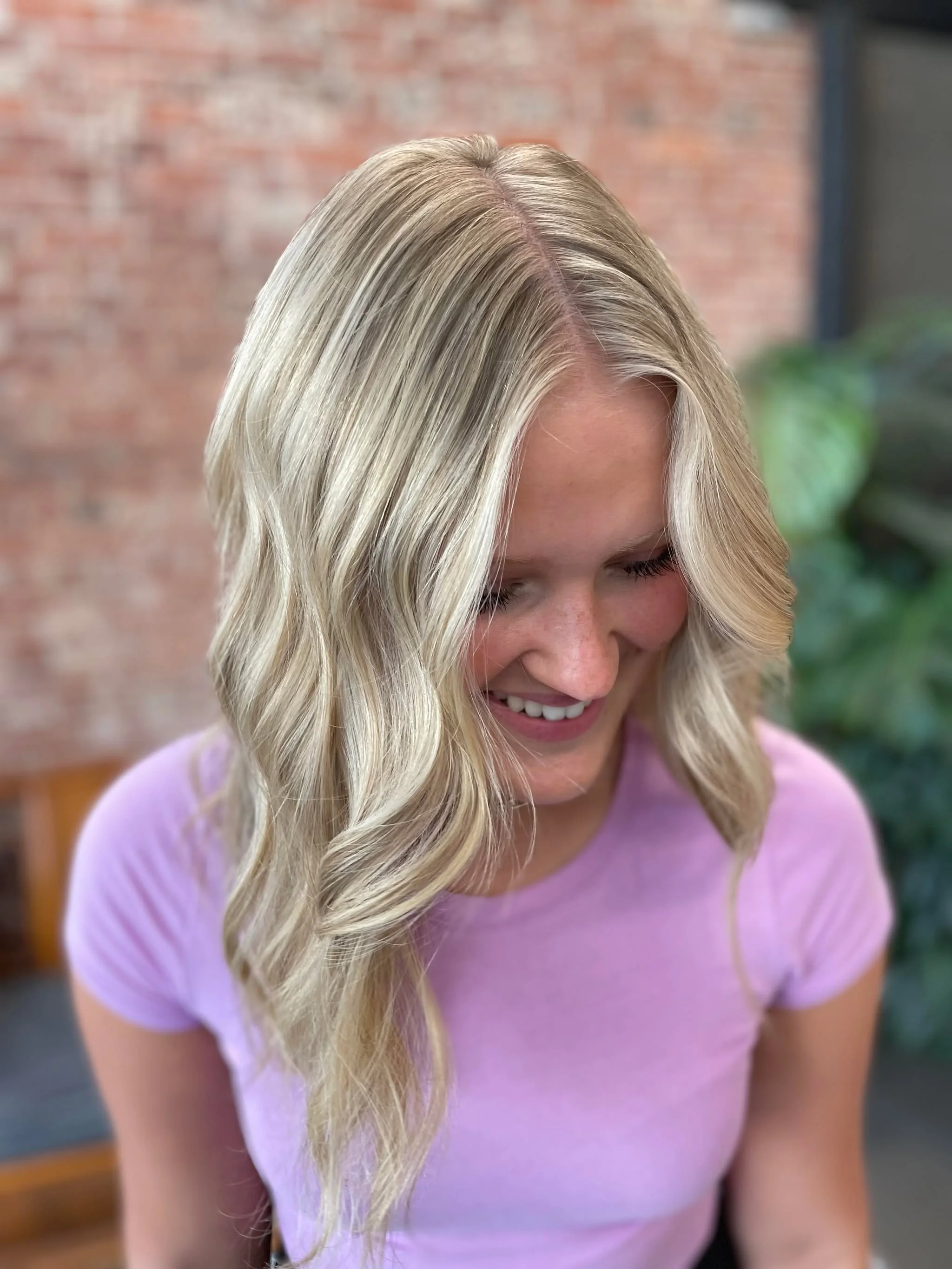 A woman with long, wavy blonde hair wearing a purple shirt, smiling and looking downward.