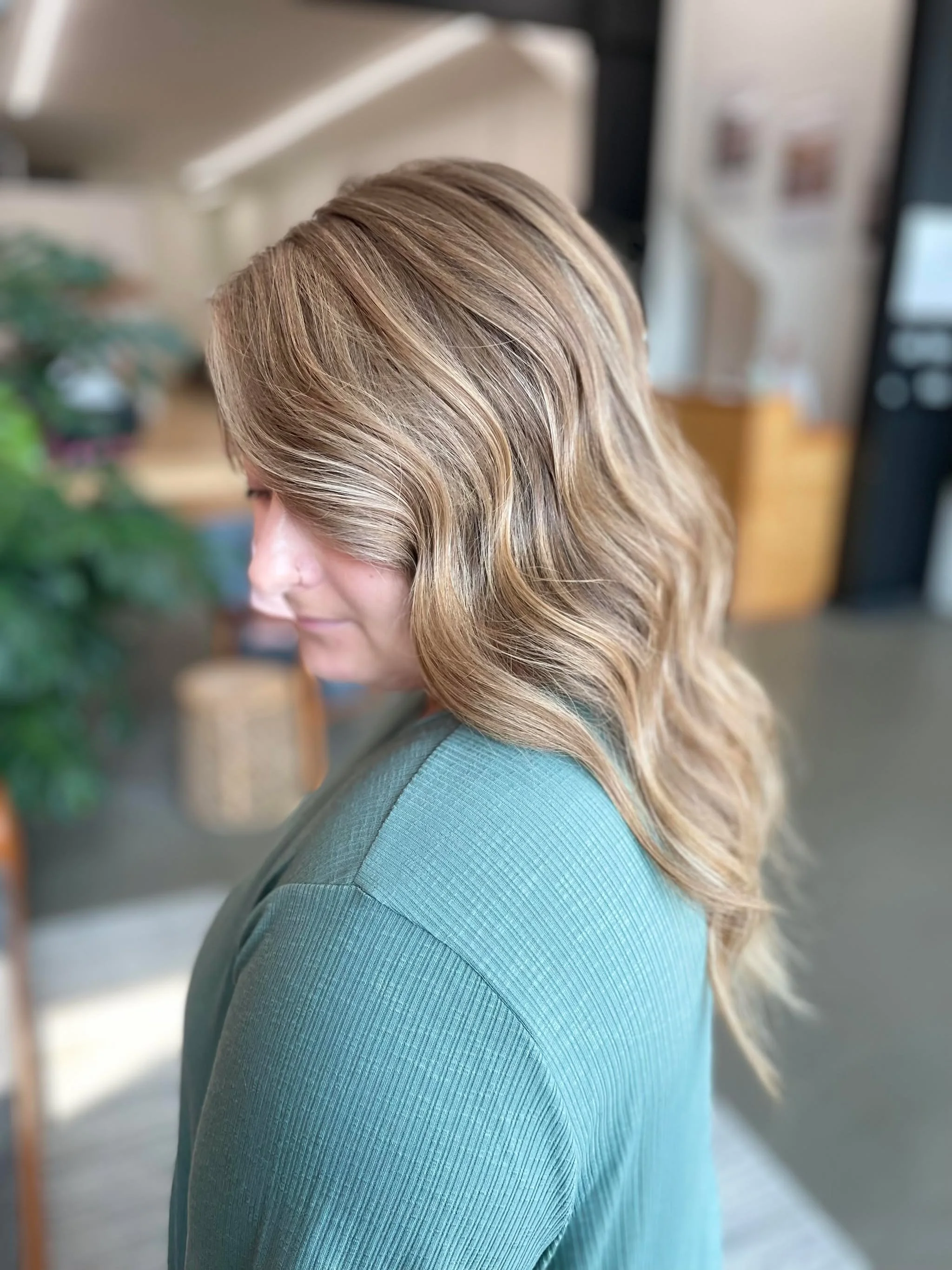 A woman with wavy blonde hair wearing a teal top standing indoors, with a blurred background of a room.