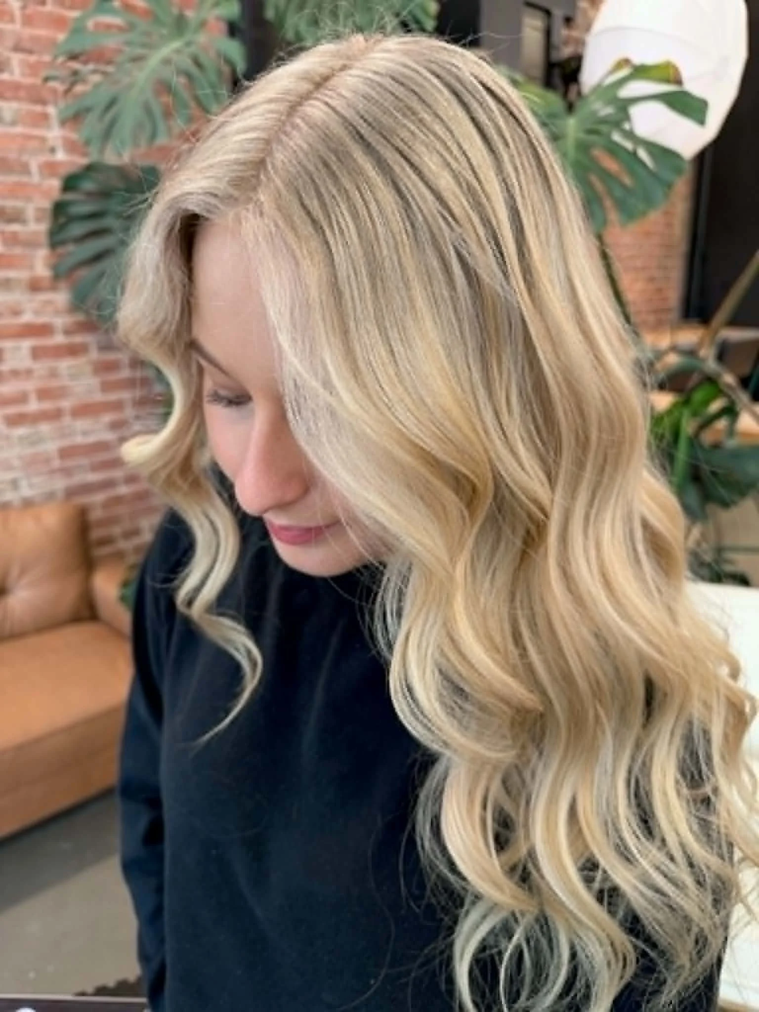 A woman with long, styled blonde hair with soft waves, wearing a dark top, in an indoor setting with exposed brick wall and large leafy green plants in the background.