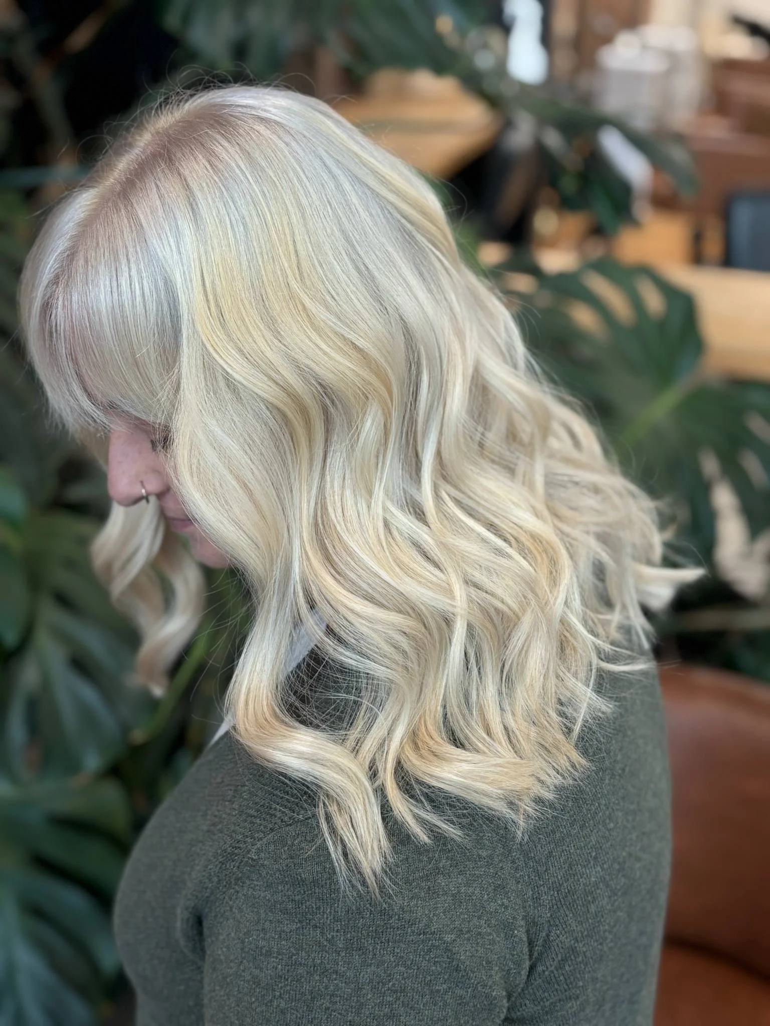 Close-up of a woman with long, wavy platinum blonde hair, seen from the side, standing indoors with greenery and wooden furniture in the background.