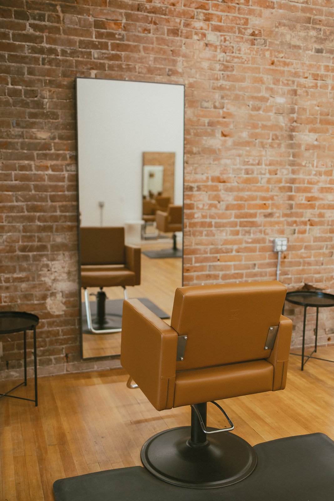Salon chair in front of a tall mirror with a brick wall background, small side tables beside the chair, and a power outlet on the wall.