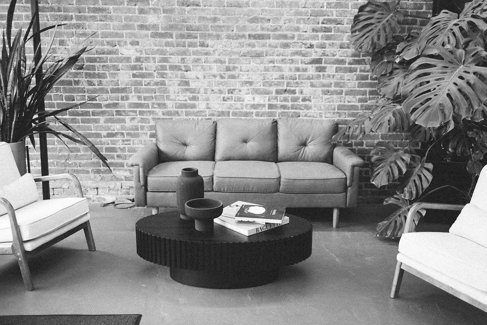 A modern salon waiting area with a brick wall, a sofa, two armchairs, a coffee table, and large potted plants.