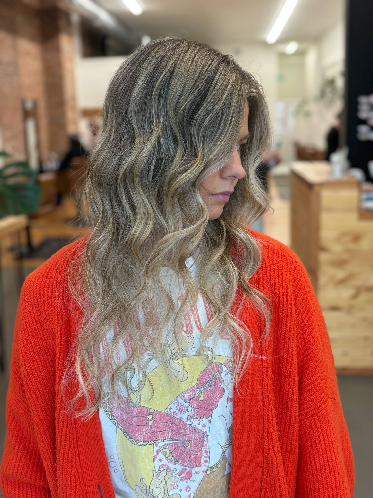 A woman with long, wavy blonde and gray hair, wearing an orange sweater and a graphic T-shirt, standing in a room with blurred background.