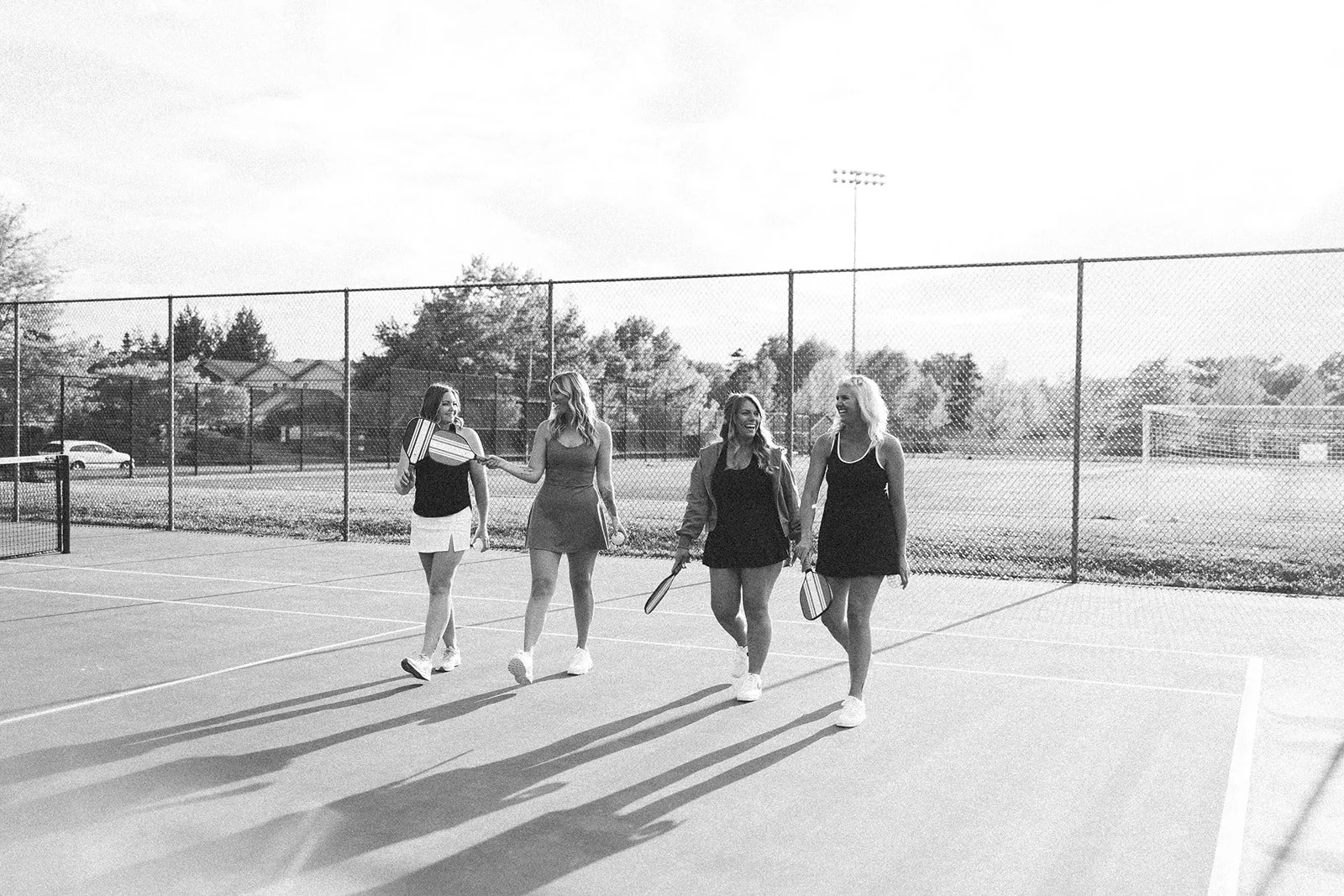 Four women walking together on a tennis court, smiling and holding tennis rackets, with a fence and trees in the background.