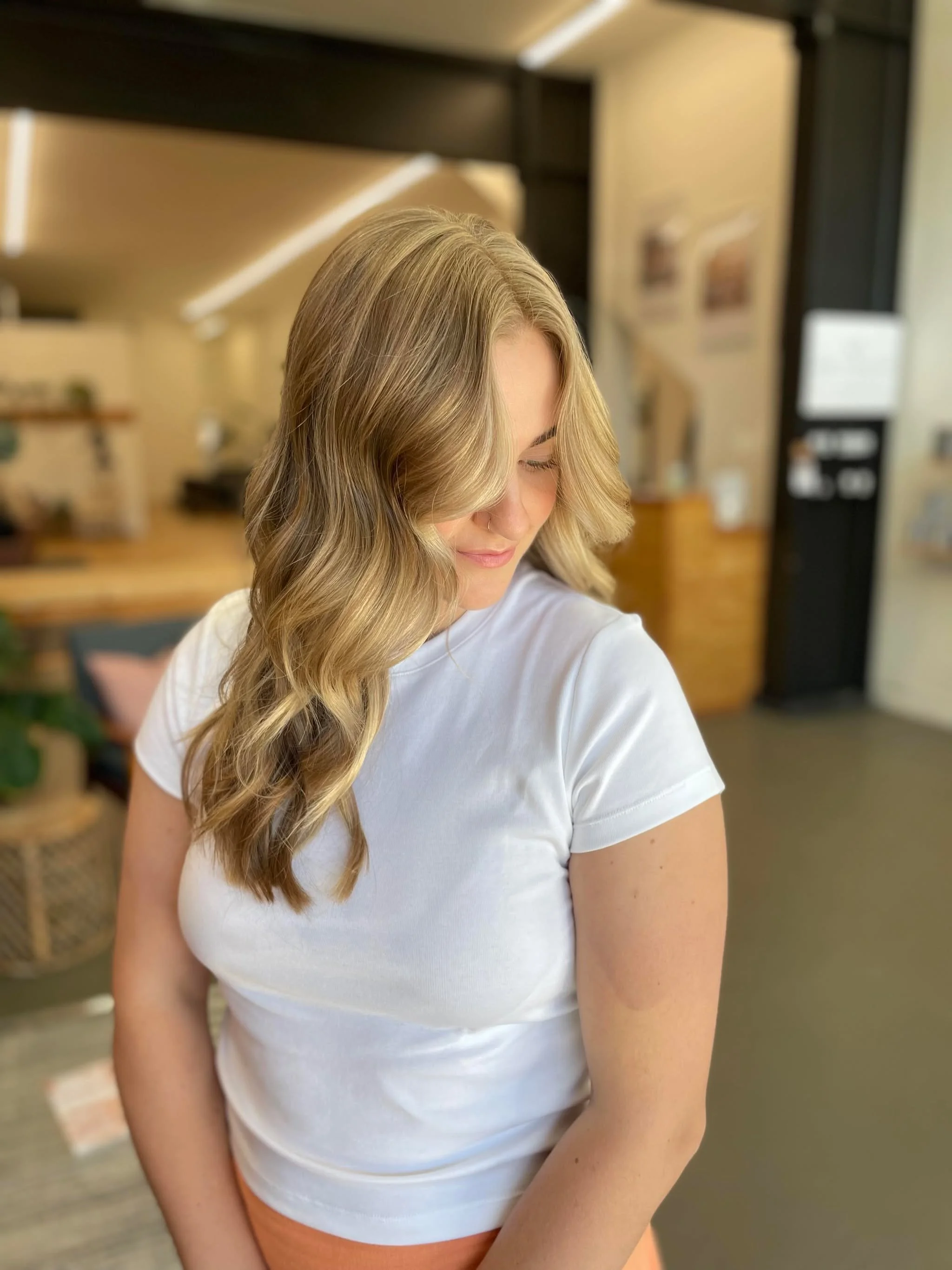 Close-up of a woman with long, blonde, wavy hair, wearing a white t-shirt, in an indoor space with blurred background.