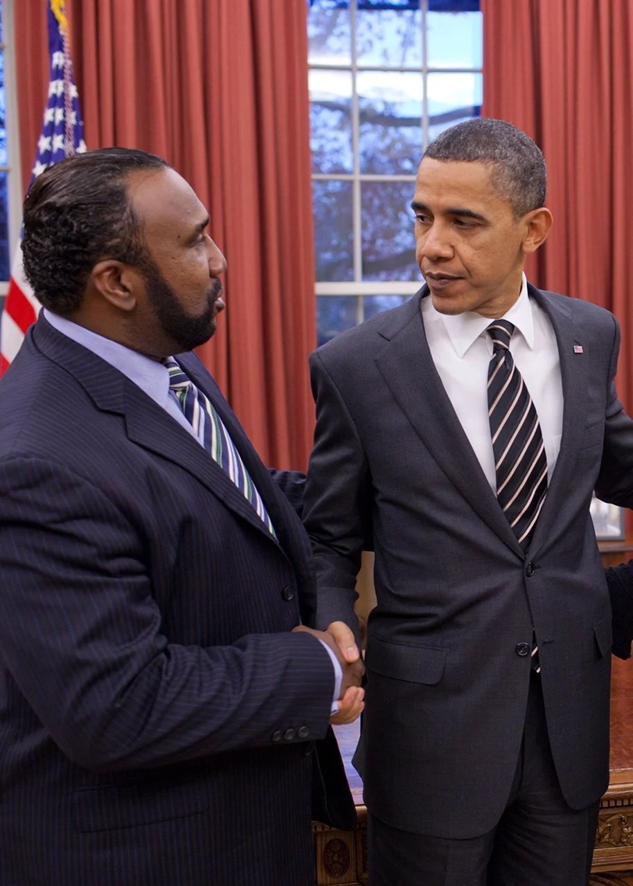 Two men in suits shaking hands in a formal setting with American flag and window with curtains in the background.