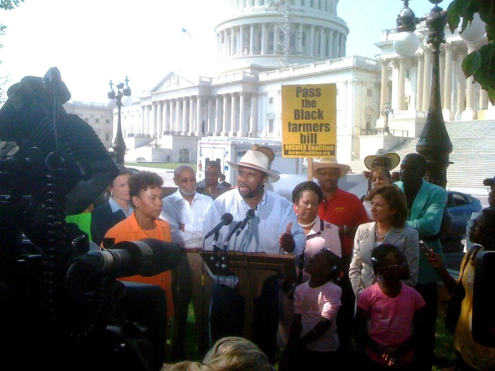 A group of people gathered outside a government building, with a man in a white shirt and hat speaking at a podium, holding a sign that reads 'Pass the Black farmers bill'.