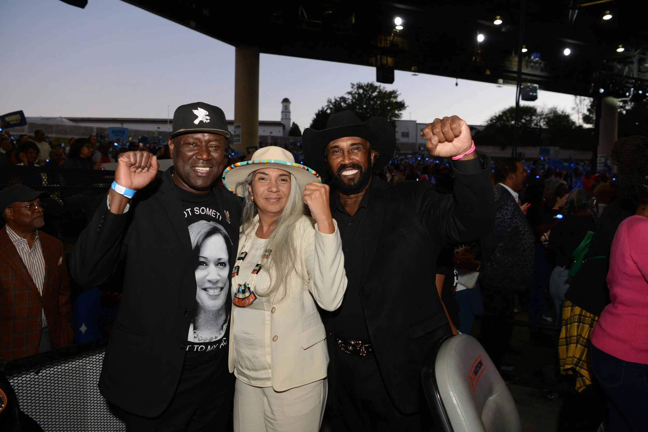 Three people posing with raised fists at an outdoor event, with a crowd and stage in the background.