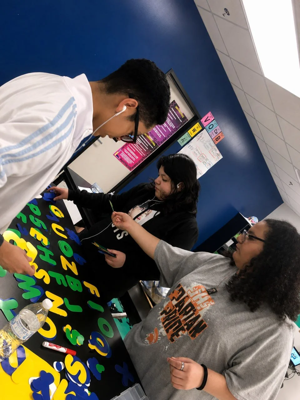Three students working on a craft project at a table covered with foam letters and shapes in green, blue, yellow, and purple.