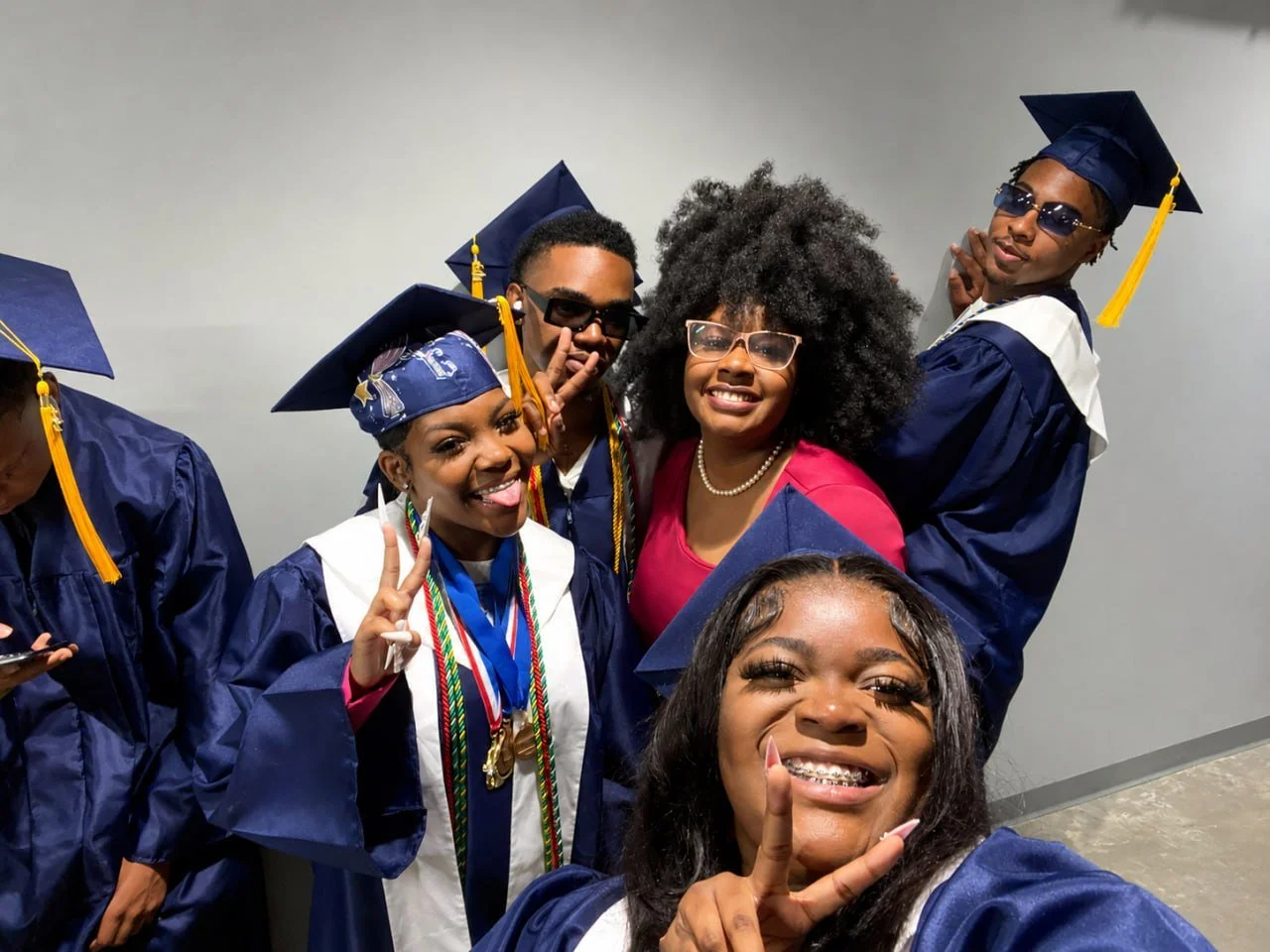 Group of five diverse young adults in graduation caps and gowns celebrating, smiling, and making peace signs at a graduation ceremony.