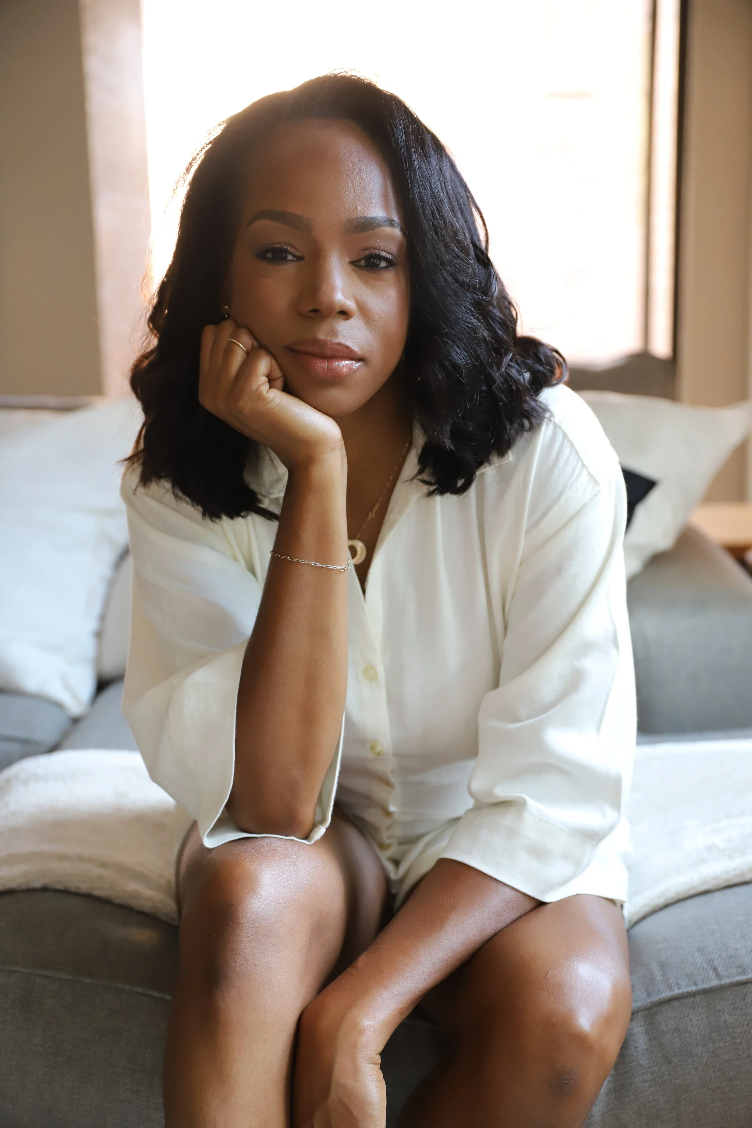 A woman with shoulder-length black hair and a light brown skin tone, sitting on a gray couch in a sunlit room, resting her chin on her hand, wearing a cream-colored shirt and minimal jewelry.