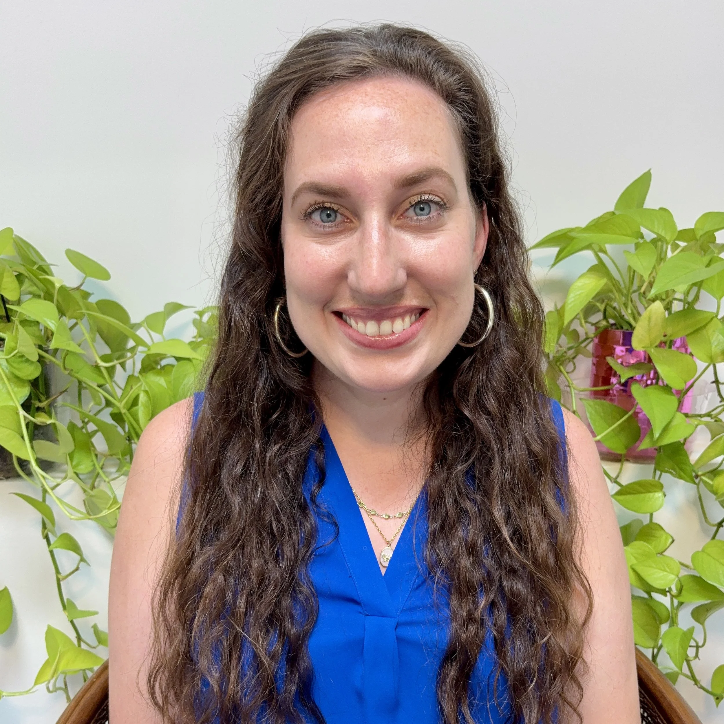 Close-up of a woman with long, curly brown hair, blue eyes, and wearing a blue top, sitting in front of green plants with a plain light background.