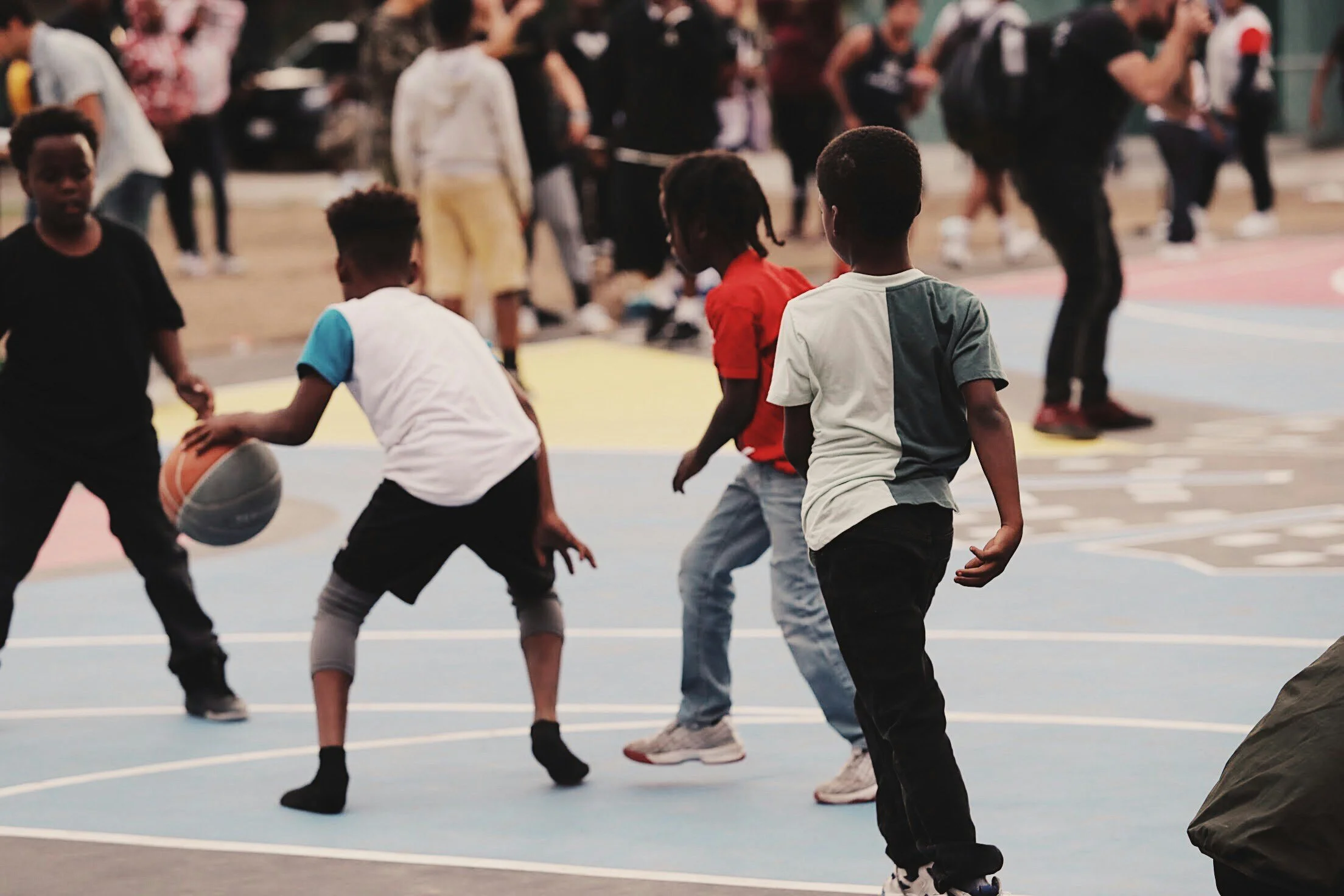 Children playing basketball on an outdoor court with many people in the background.