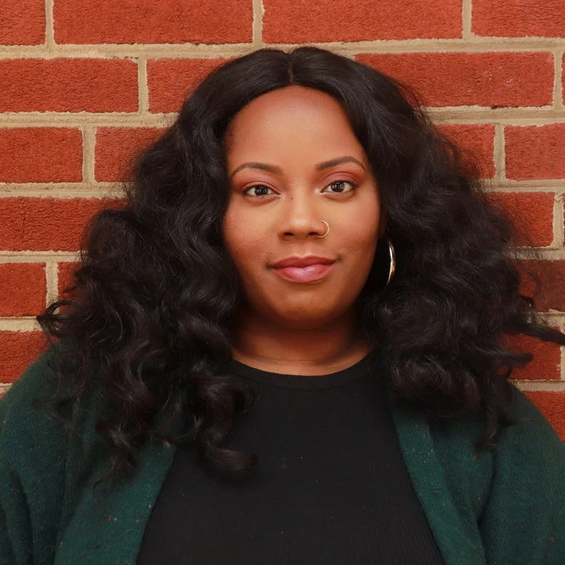Portrait of a woman with long, curly black hair, a nose ring, and hoop earrings standing against a red brick wall.