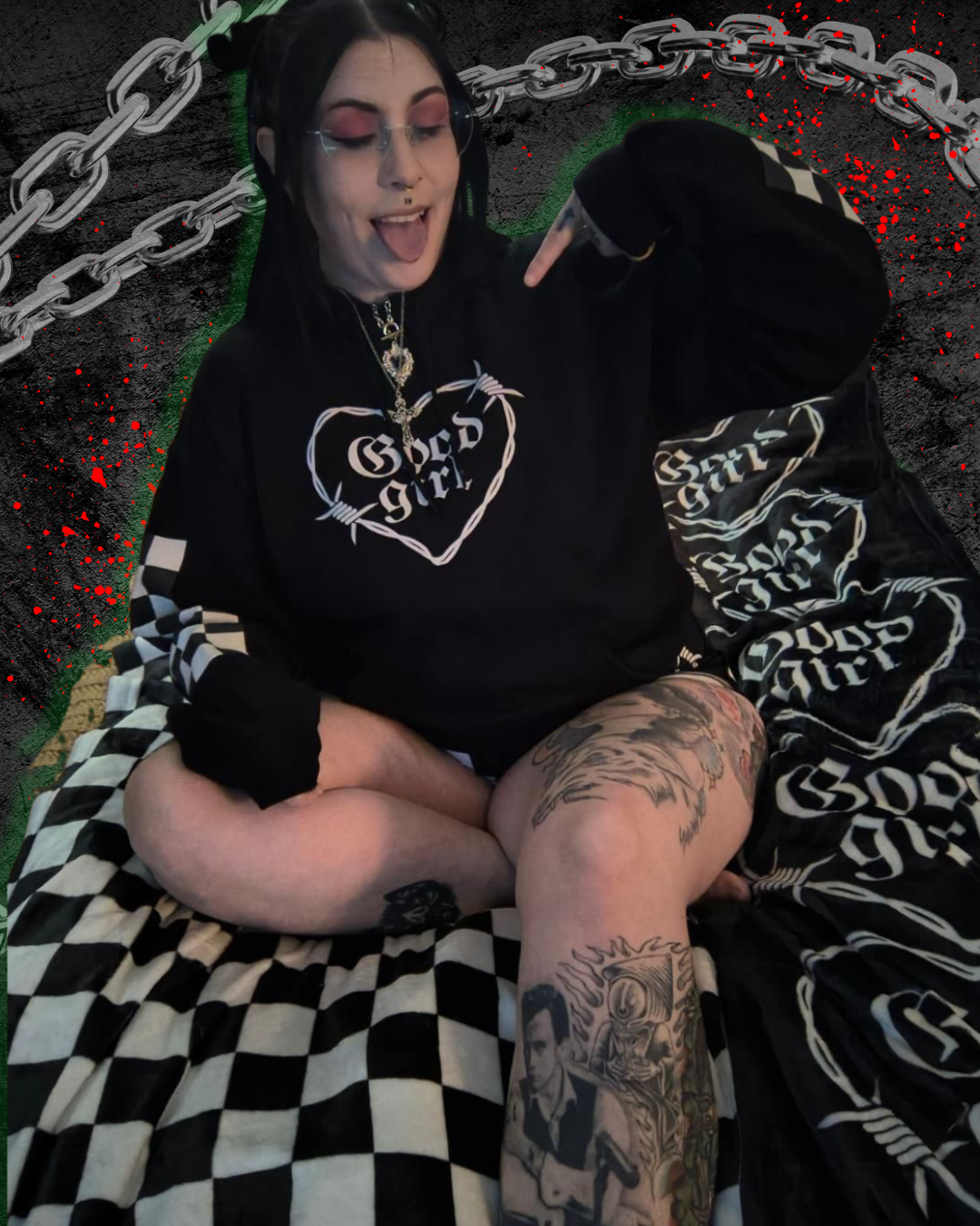 A person with black hair and tattoos on their leg, wearing glasses, a black hoodie with white lettering, and checkered shorts, sitting cross-legged on a black-and-white checkered blanket, making a gesture with their middle finger, with a dark background featuring a chain graphic and red splatters.