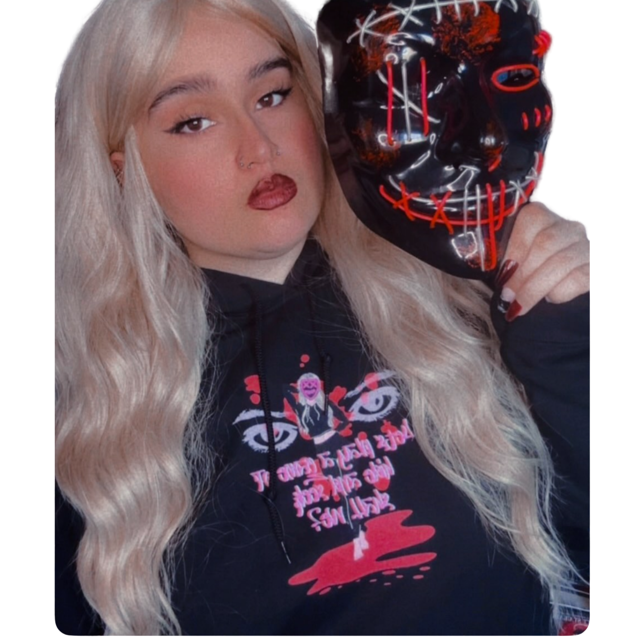 A person with long blonde hair and makeup holding a black mask with red and white stitching design.
