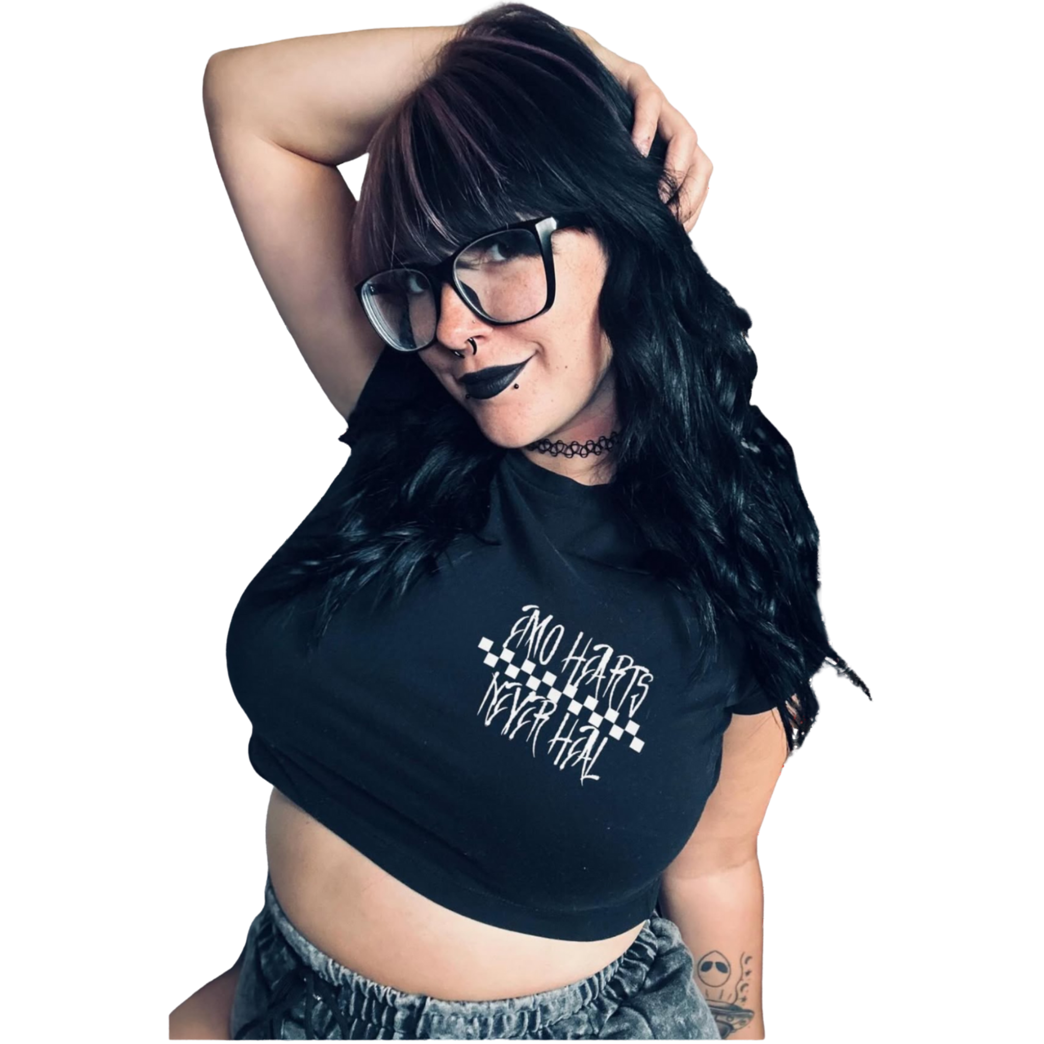 A woman with black hair, glasses, and dark lipstick, wearing a black t-shirt with white text, standing with her hand on her head, showing a tattoo on her arm.