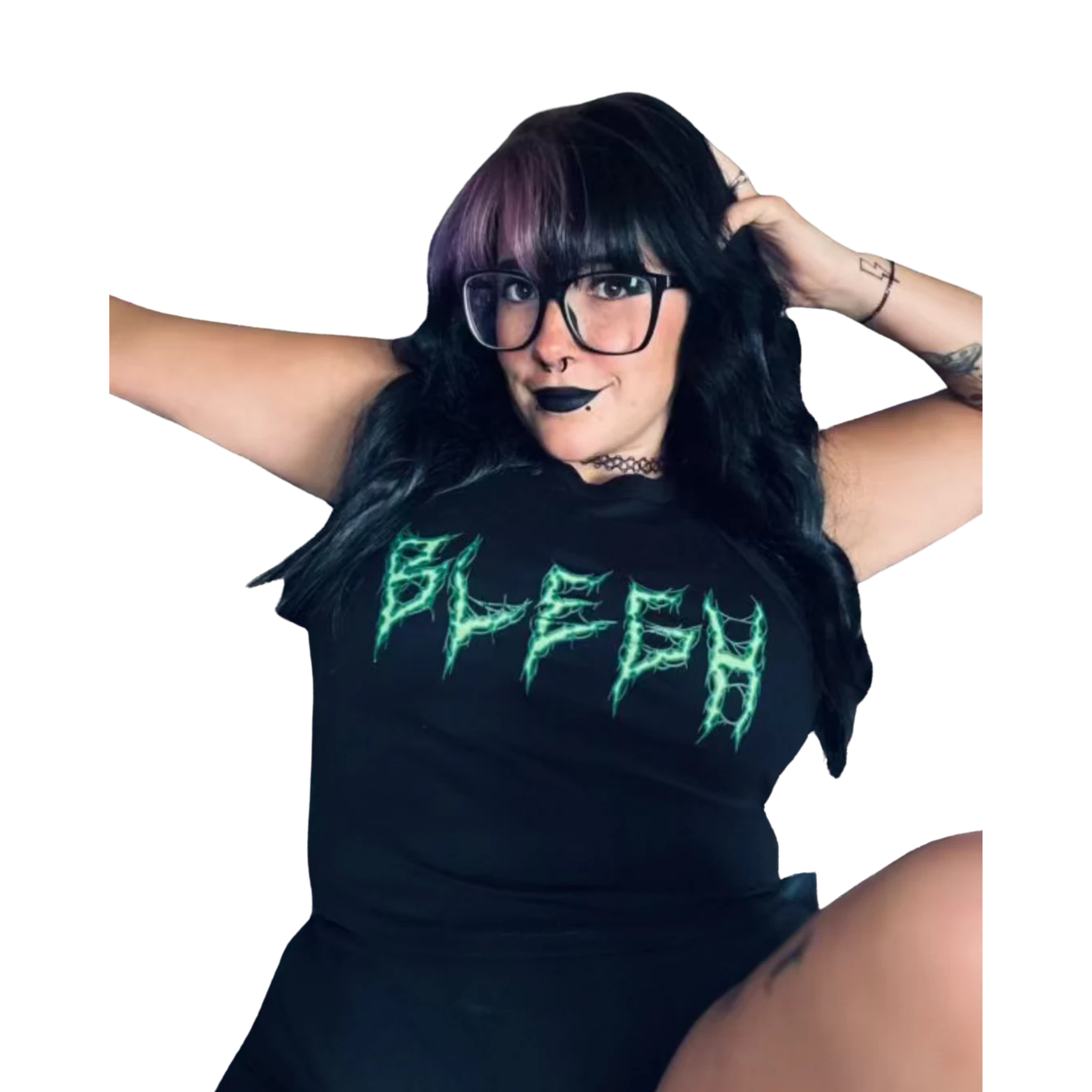A woman with black hair, glasses, and dark lipstick poses with her hands in her hair, wearing a black t-shirt with green lettering. The background is plain black.