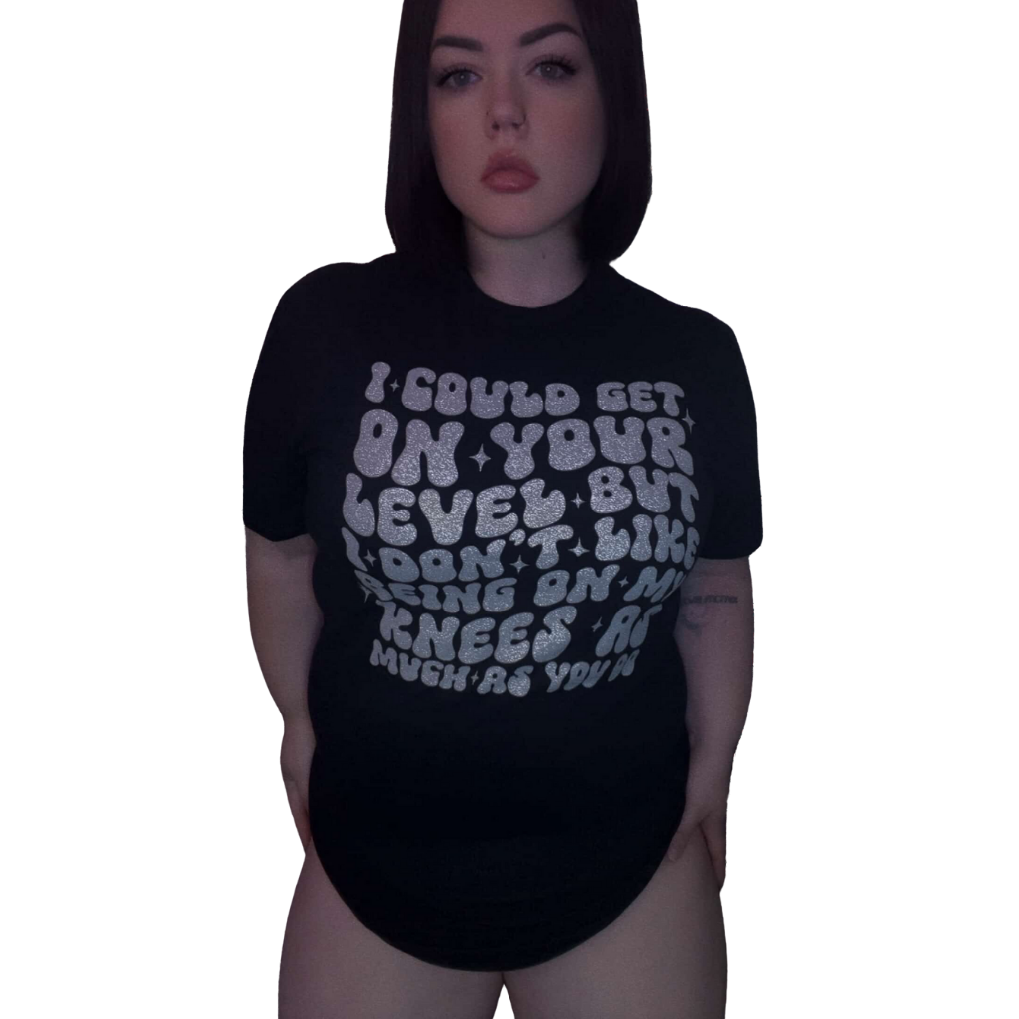 A woman with dark shoulder-length hair and light skin is wearing a black top with white glittery text and black underwear, standing against a black background.