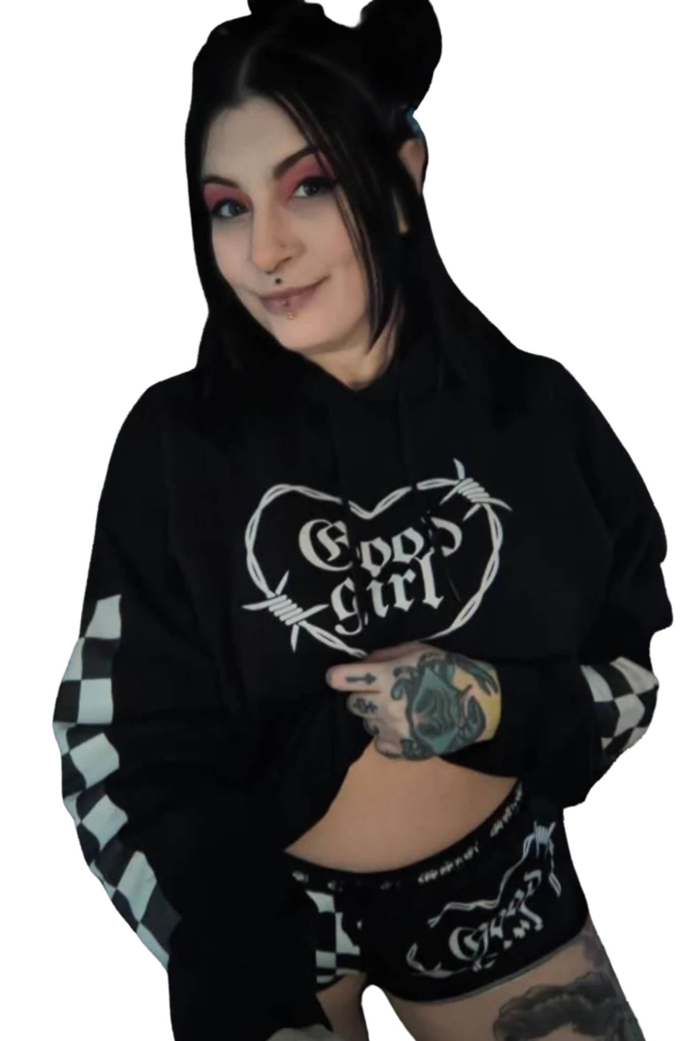 A woman with black hair and tattoos on her arm and thigh, wearing a black hoodie with white checkered sleeves, smiling and pointing to her stomach.