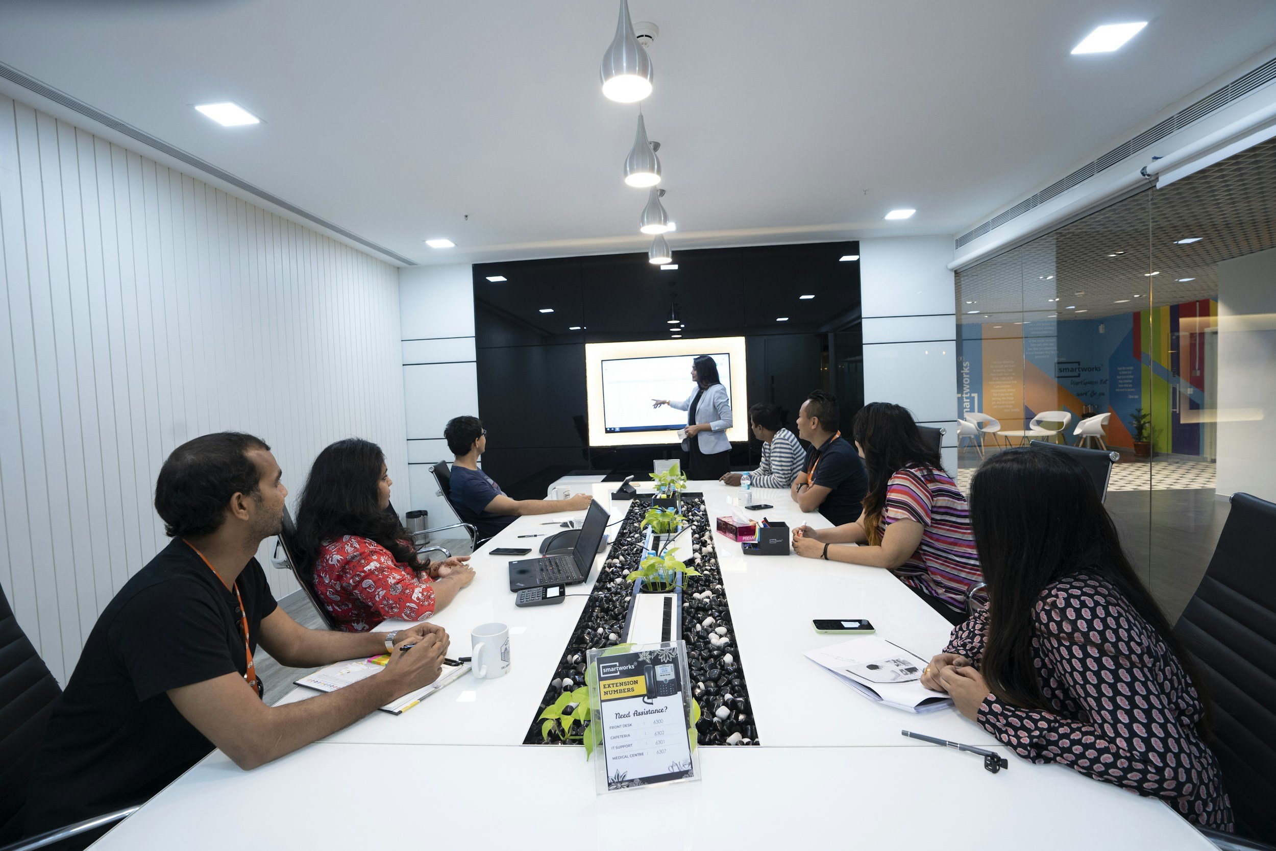 A business meeting in a modern conference room with a diverse group of people sitting around a large white table, listening to a woman in a blazer giving a presentation on a large screen. The room has contemporary lighting, glass walls, and a colorful mural in the background.