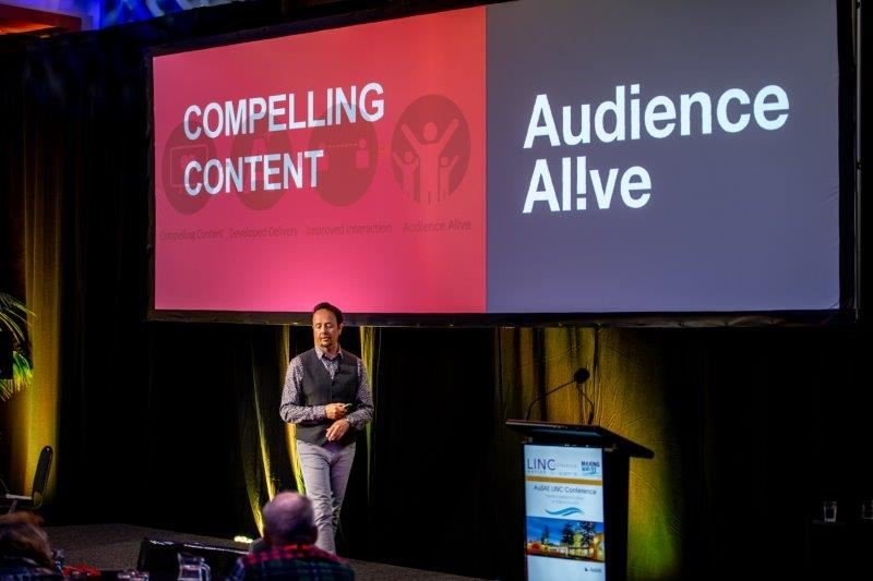 A speaker on stage at a conference, with large screen behind him displaying a slide that reads 'Compelling Content' and 'Audience Alive.'