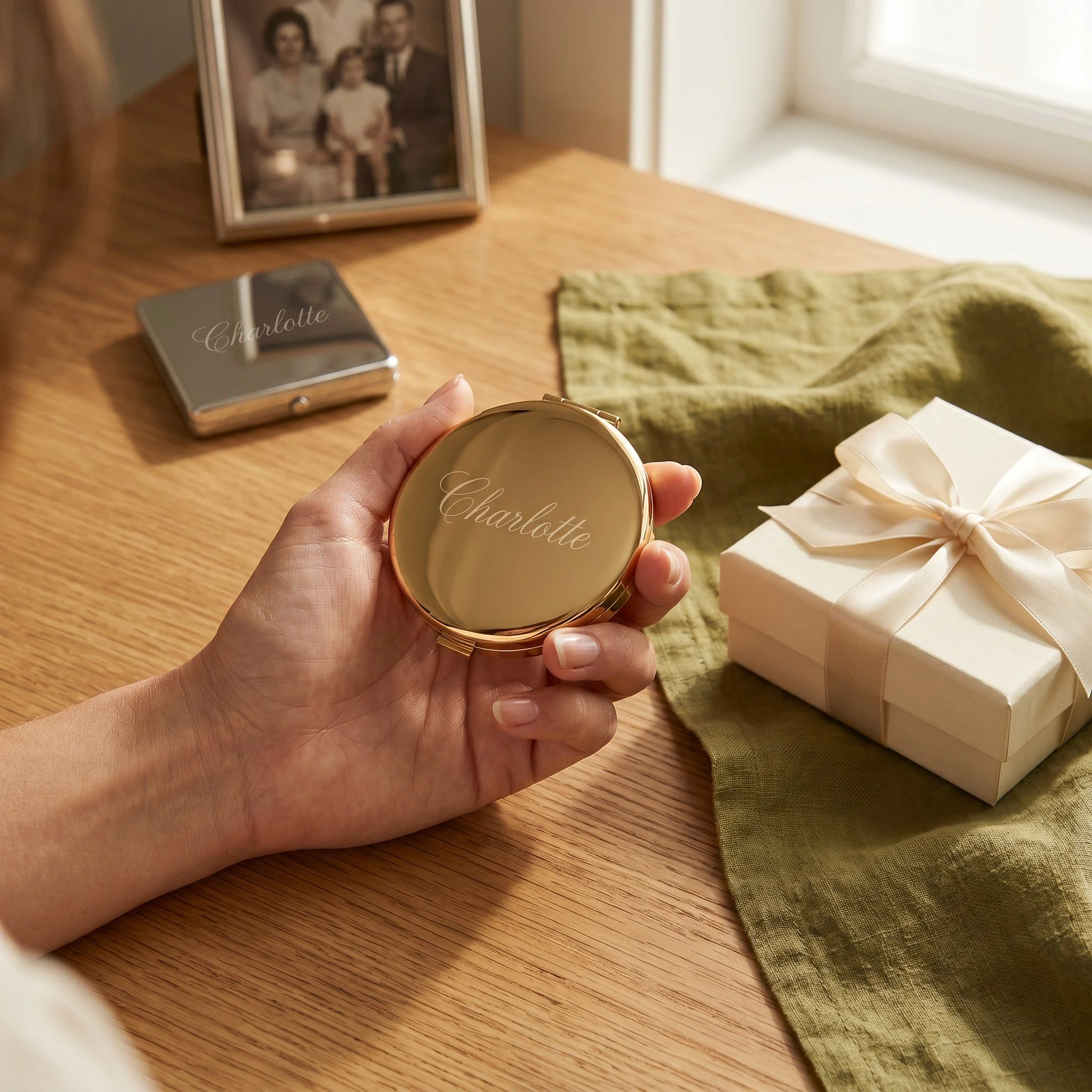Close-up of a hand holding a gold-colored round box with the name Charlotte written on the lid, on a wooden surface. In the background, there is a framed family photograph, a black and silver cardholder, a wrapped gift box with a ribbon, a green fabric, and a partially visible window letting light into the room.