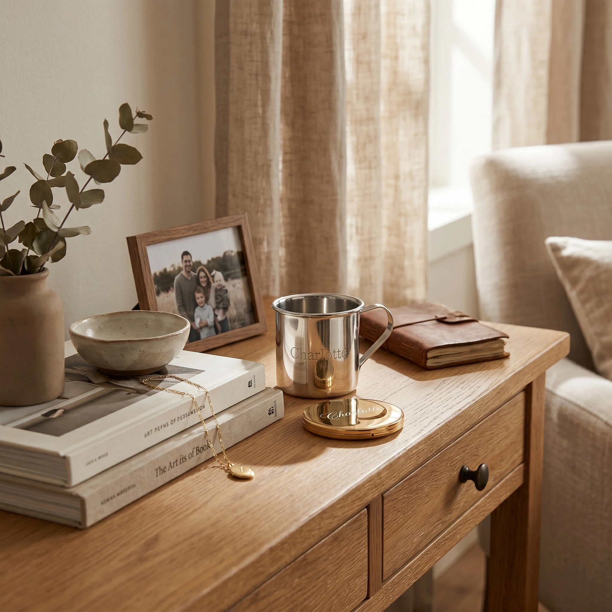 A wooden table with a framed family photo, books, a ceramic bowl, a metal mug labeled 'Charlotte', a jewelry box, a potted plant, a gold necklace, and a round container with a lid. In the background, a window with beige curtains.