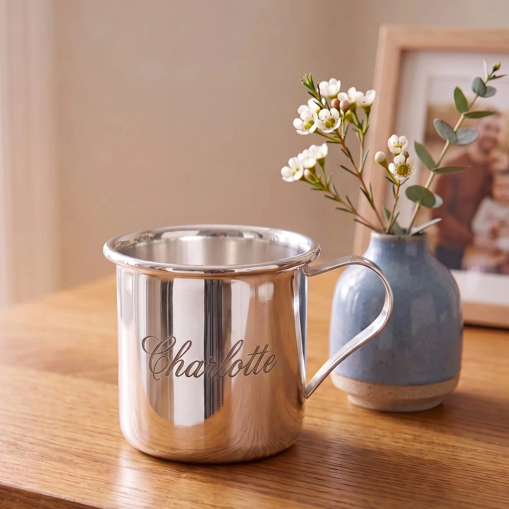 A silver mug with the name 'Charlotte' engraved on it, placed on a wooden surface, with a blue vase containing white flowers and green foliage in the background.