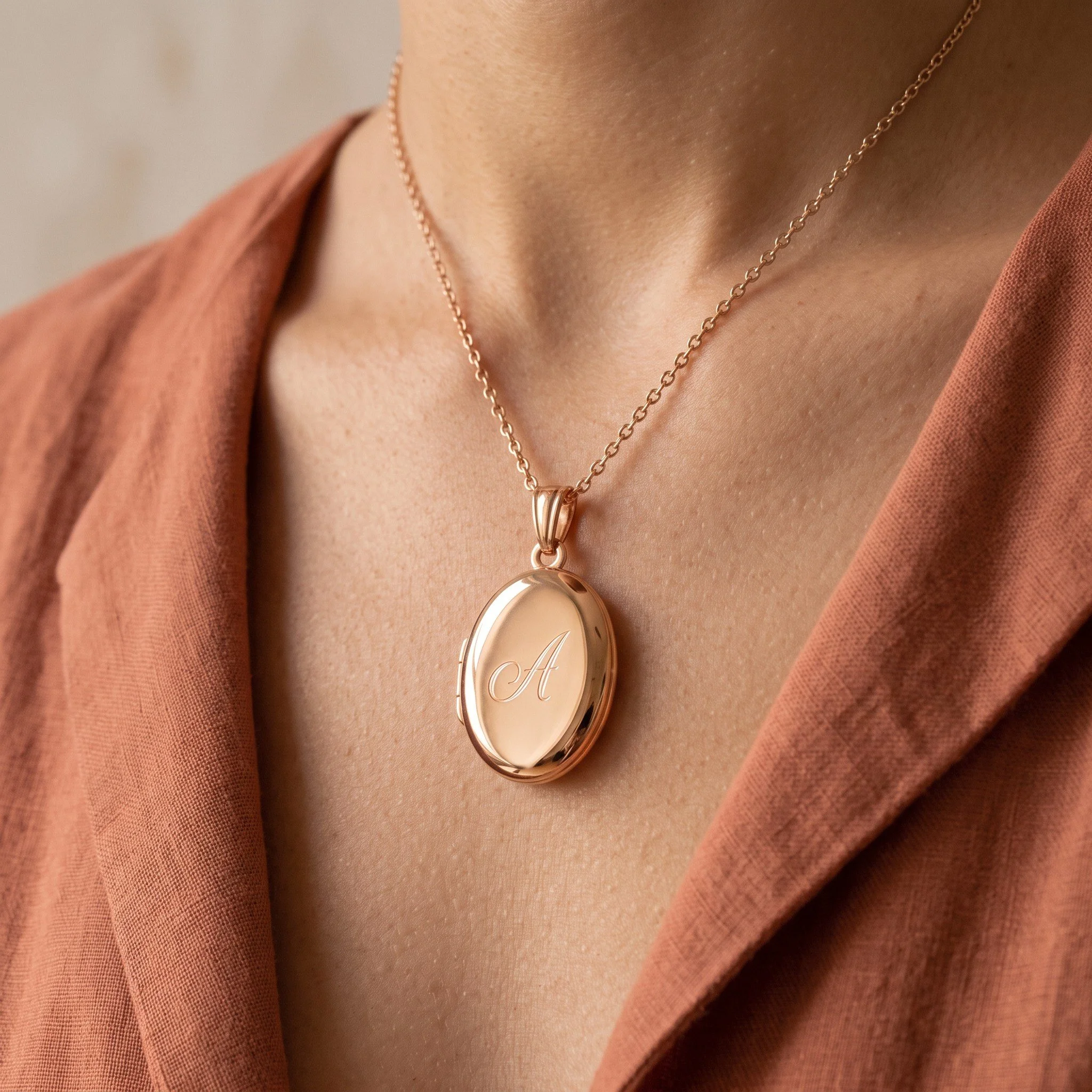 Close-up of a person's neck wearing a delicate gold chain necklace with a pendant engraved with the letter 'A', against a rust-colored fabric background.