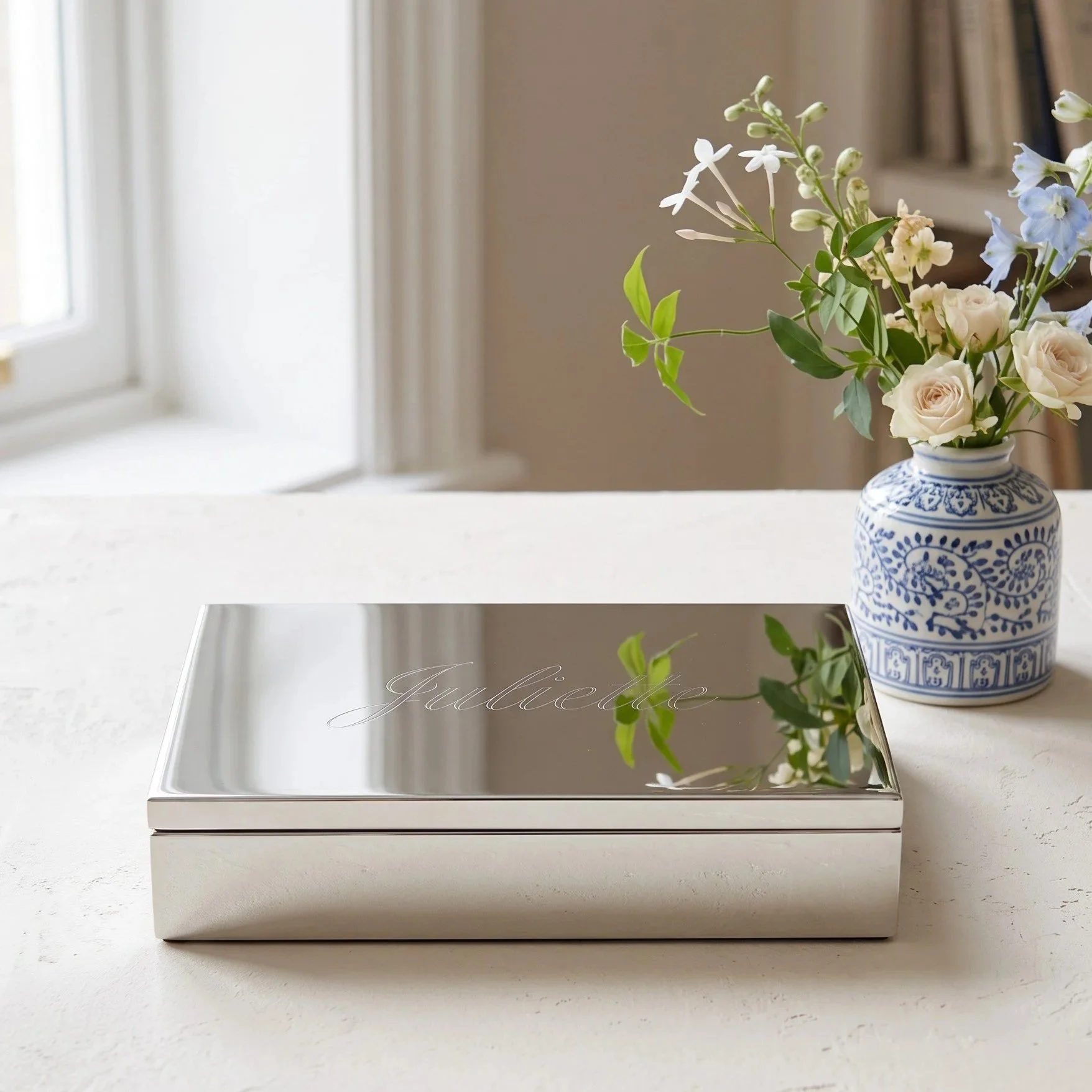 A white flower vase with a blue decorative pattern, containing white roses and other small white and blue flowers, on a white surface with a mirrored box in front that has the word 'Guillette' written on it, reflection of the flowers and vase visible on the mirror.