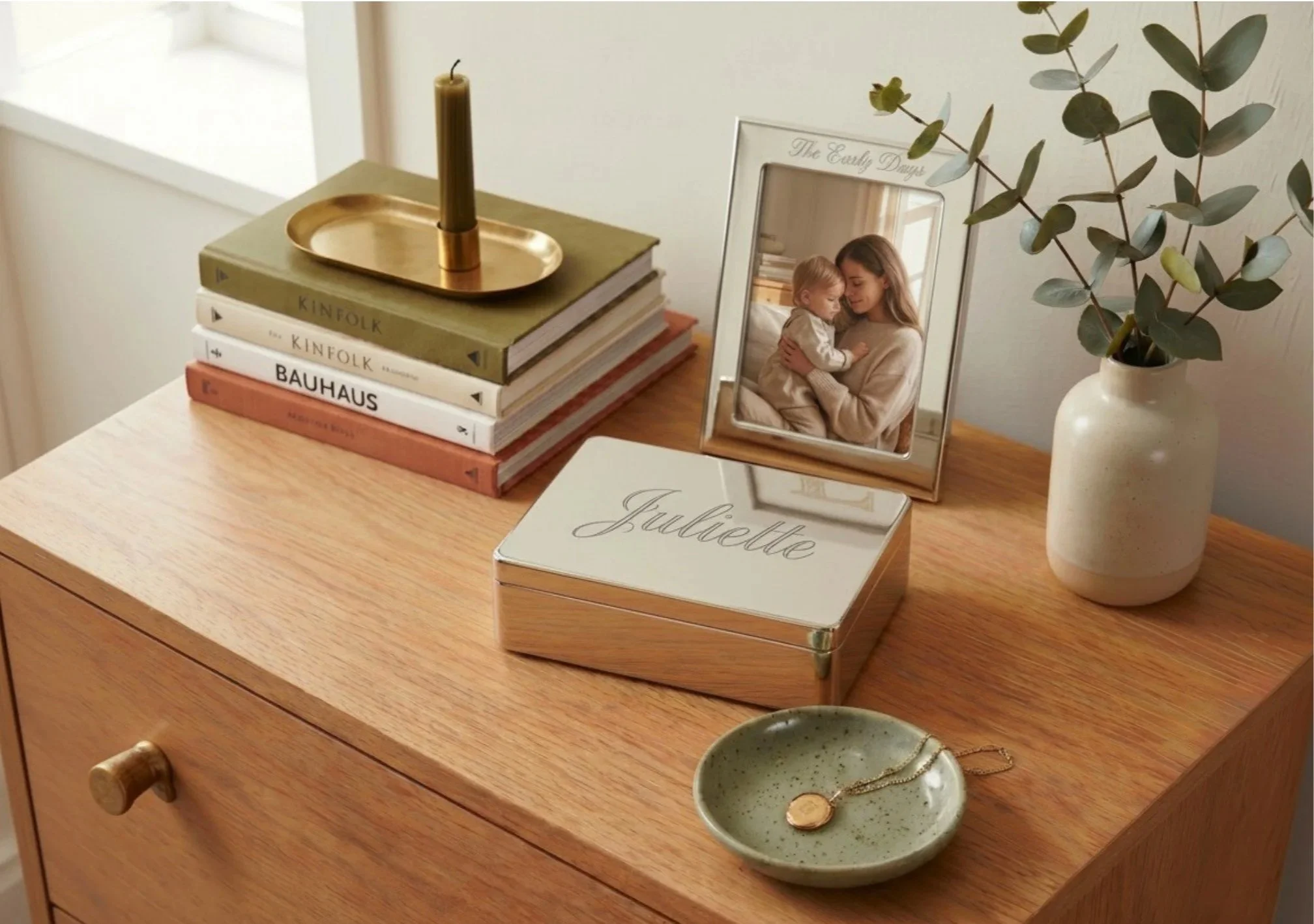 A wooden dresser with a stack of books, a framed photograph, a white vase with green foliage, a jewelry dish with a gold chain and ring, and a decorative box with the name 'Juliette' written on it.