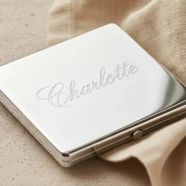A closed mirrored compact with the name Charlotte engraved in cursive on the lid, resting on beige fabric.