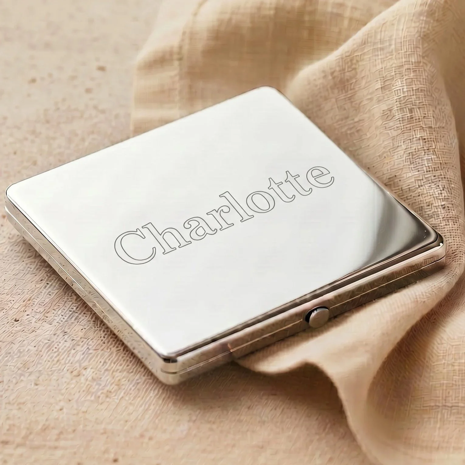 A silver iPhone with the word 'Charlotte' engraved on the back, resting on a beige textured surface with a beige cloth nearby.