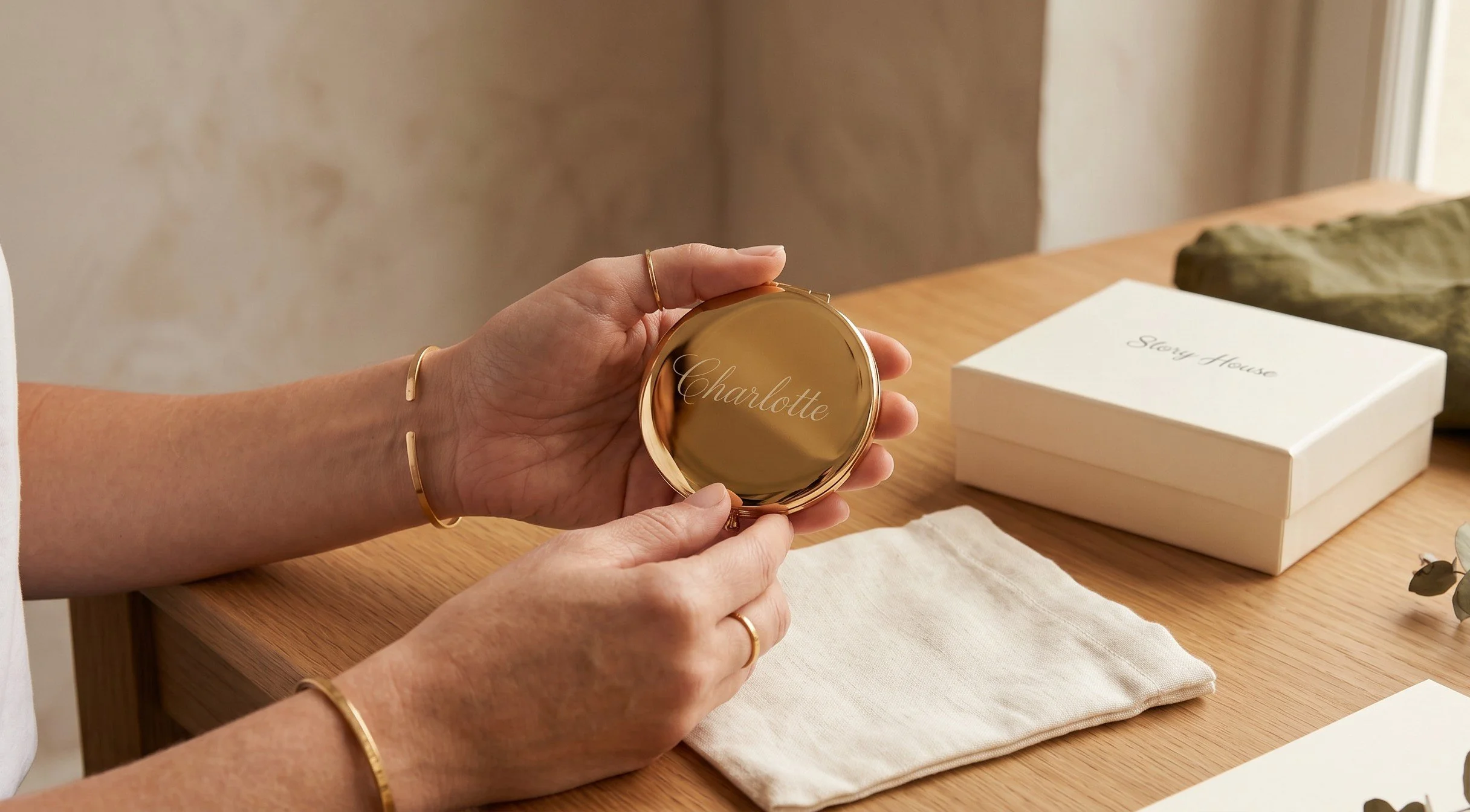 Person holding a round compact mirror with the name 'Charlotte' engraved on the lid, sitting at a wooden table with a white box labeled 'Story House' nearby.