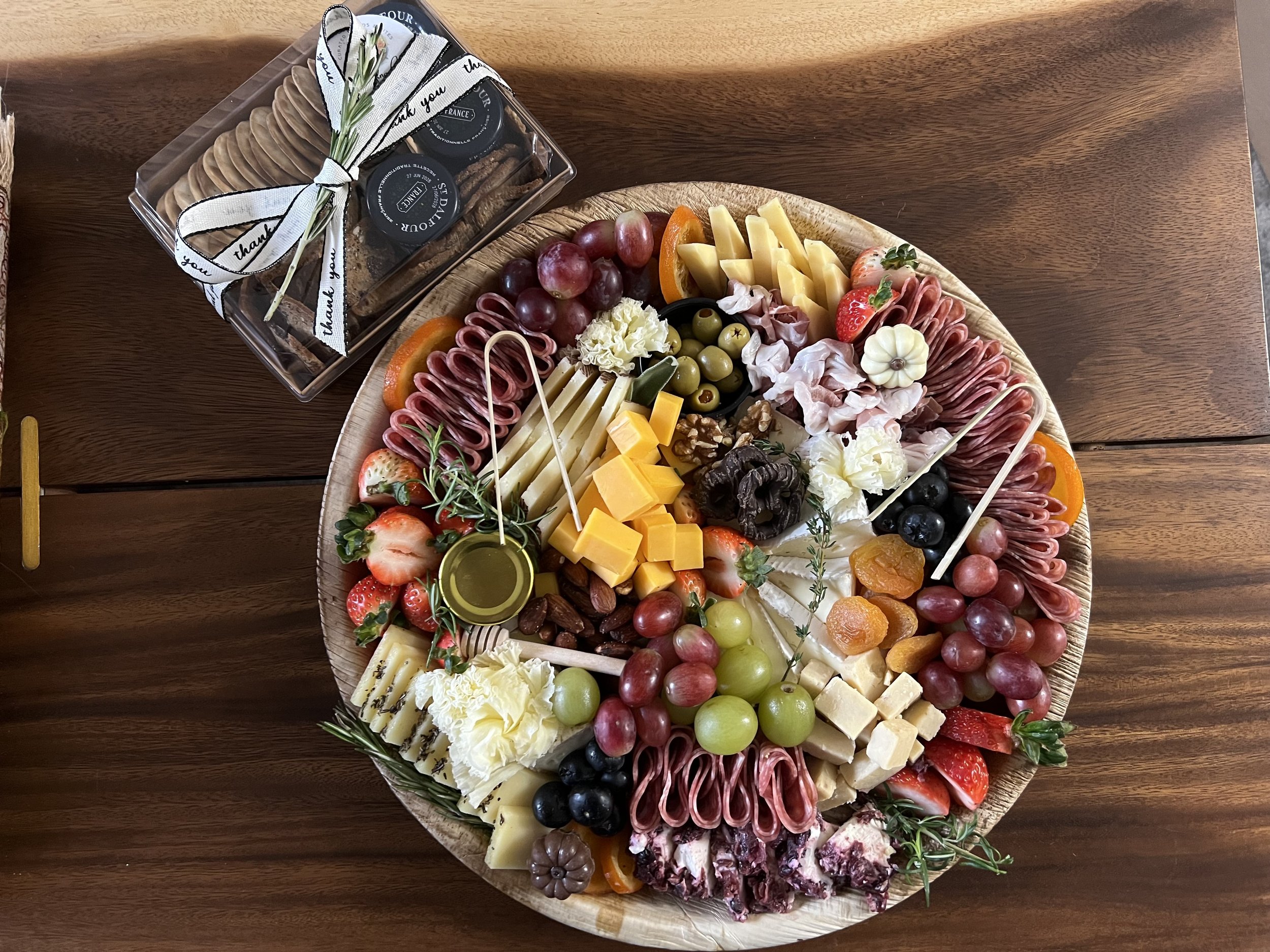 A large wooden platter holds an assortment of cheeses, grapes, strawberries, sliced oranges, and various cured meats, garnished with herbs and edible flowers. A smaller clear box contains chocolate biscuits wrapped with a bow and labeled 'Thank you' and 'thank you' ribbon.