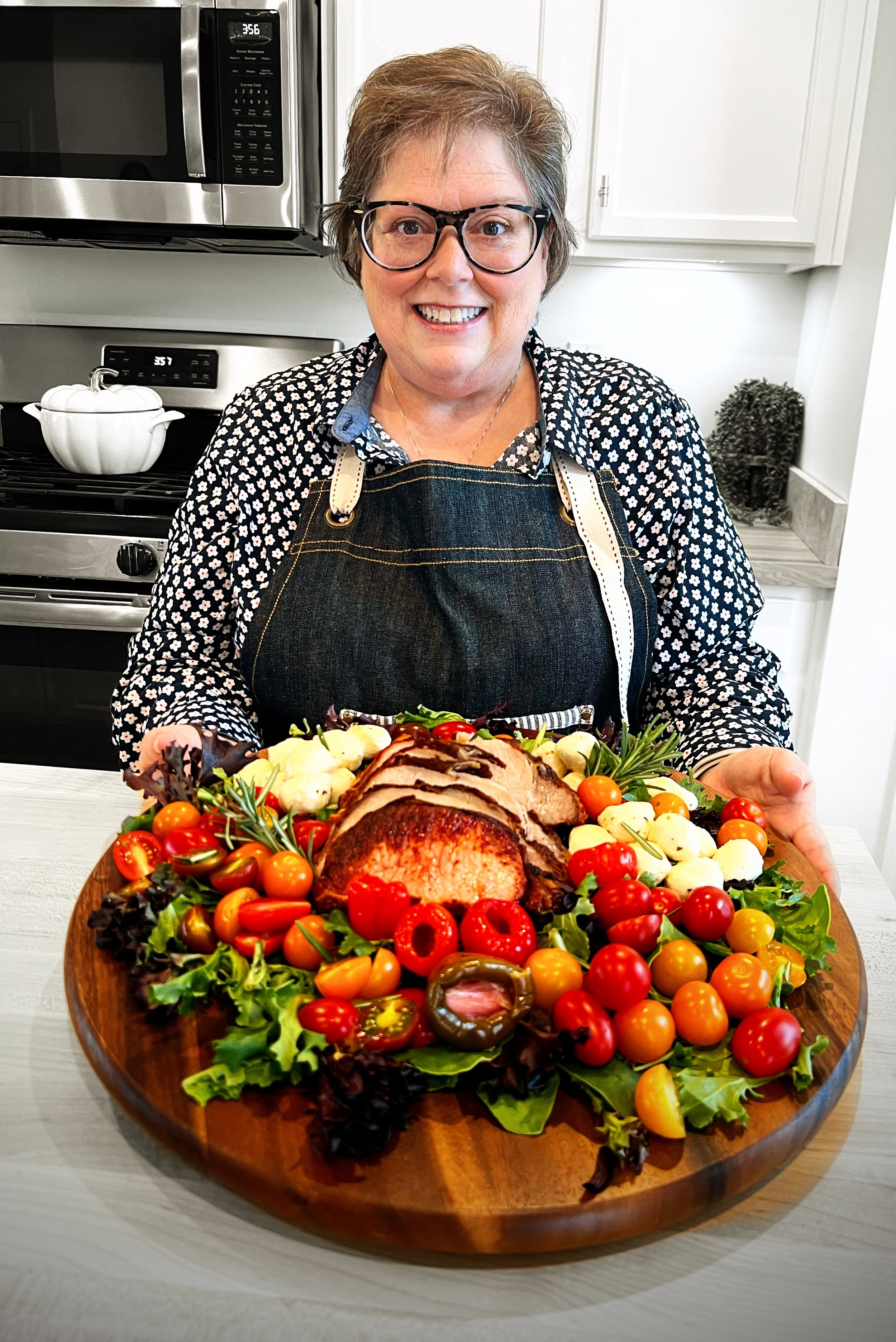 A woman with short hair, glasses, and a floral shirt, smiling and holding a large wooden platter of sliced roast beef surrounded by cherry tomatoes, greens, and vegetables in a kitchen.