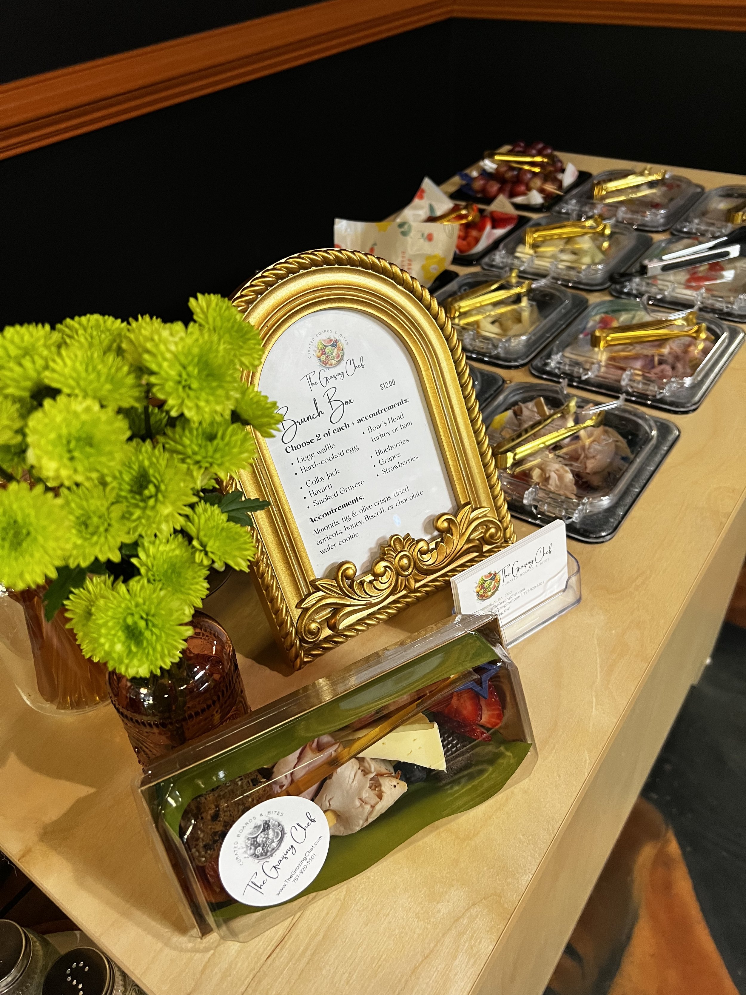 A brunch buffet offered on a mobile charcuterie cart featuring small containers of meat, cheese, and fruit, with a decorative gold-framed menu and a green plant in a small amber vase in the foreground.