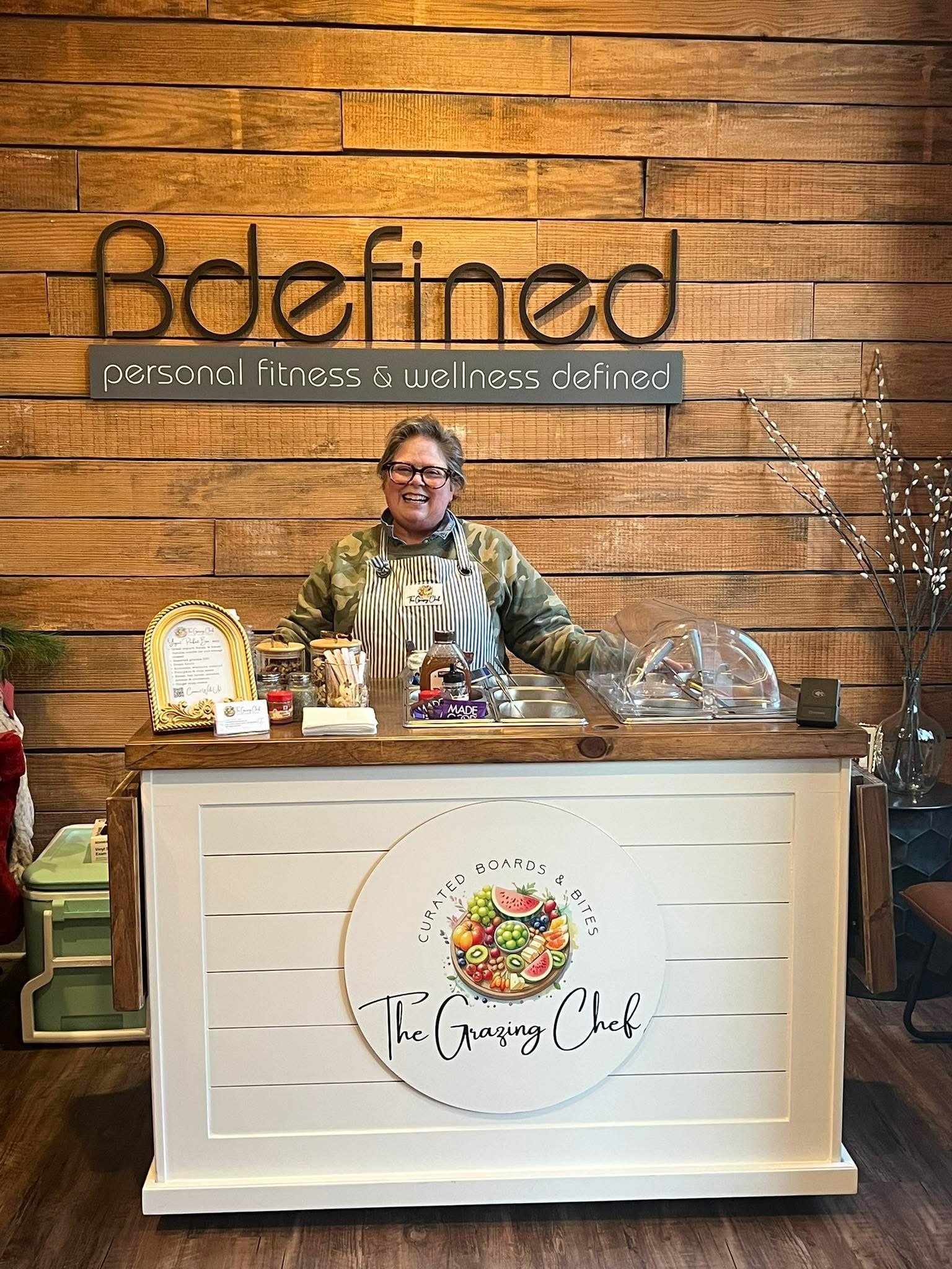 Chef standing behind a white counter with a logo that reads "The Grazing Chef." She is wearing glasses, a striped apron, and a camouflage shirt, with a wooden wall in the background bearing a sign reading "Bdefin personal fitness & wellness defined."