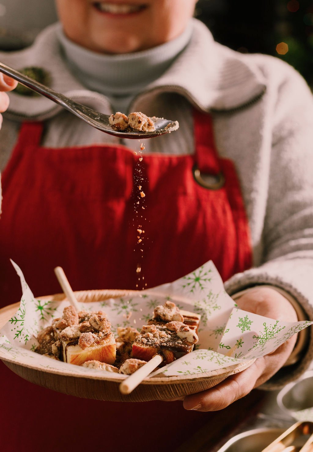 Chef Lorene, in a gray sweater and red apron, is holding a wooden plate with several decorated desserts covered in powdered sugar and crushed topping, while sprinkling additional topping onto them.