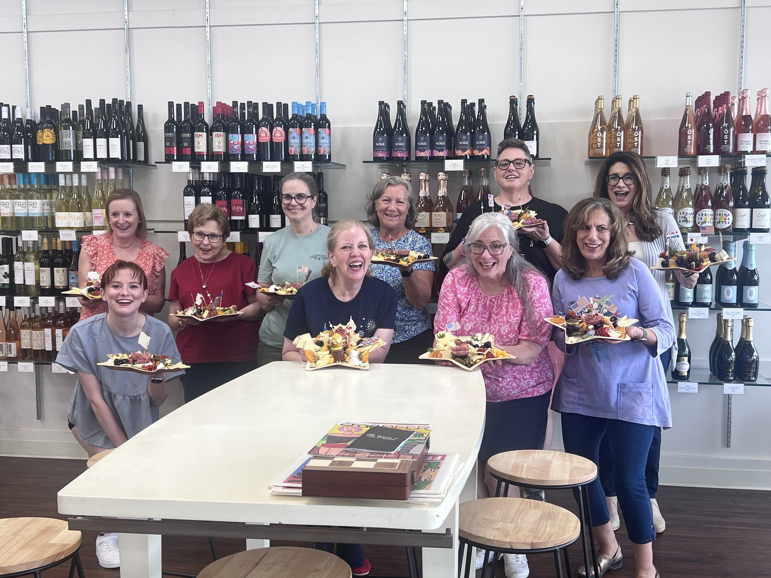 A group of women and a girl, smiling and holding charcuterie plates they designed in a workshop, standing in front of shelves stocked with wine and other beverages in a store.