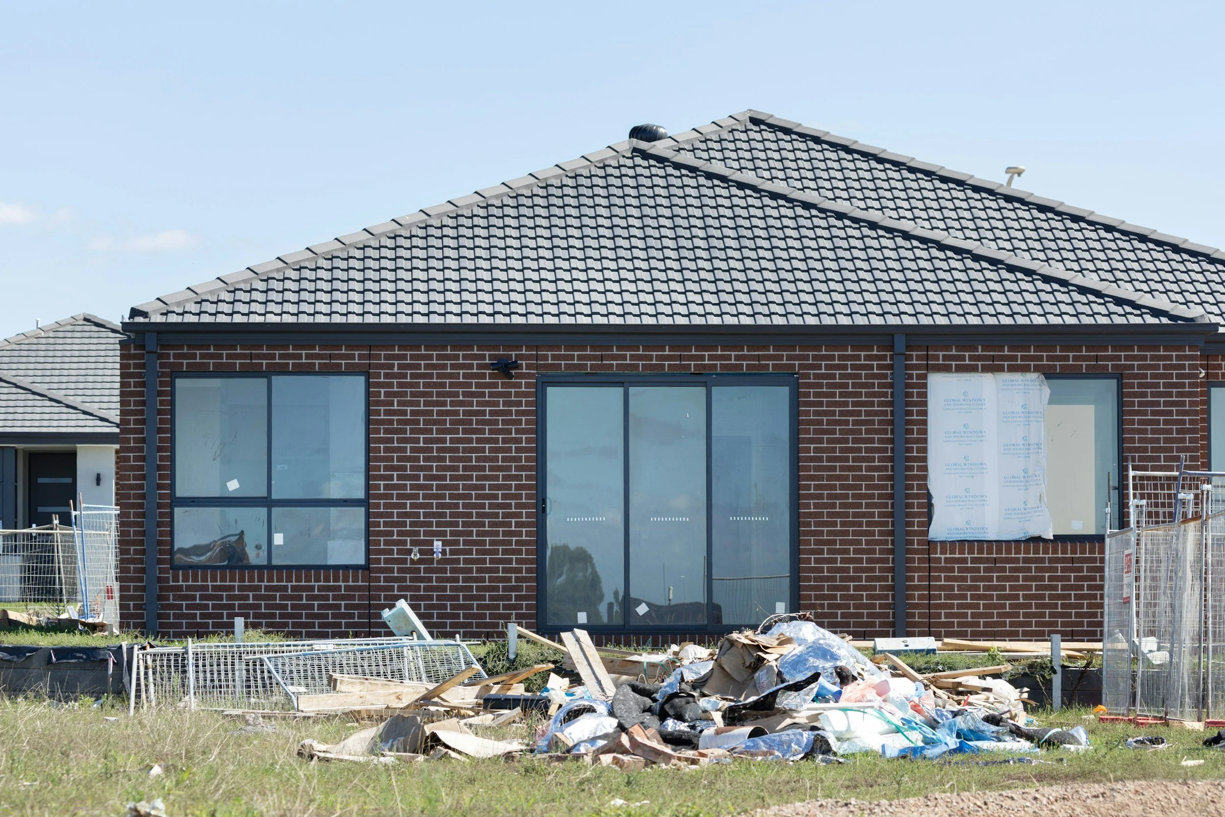 Newly built brick house under construction with large glass doors and windows, surrounded by construction debris and fencing, under a clear blue sky.